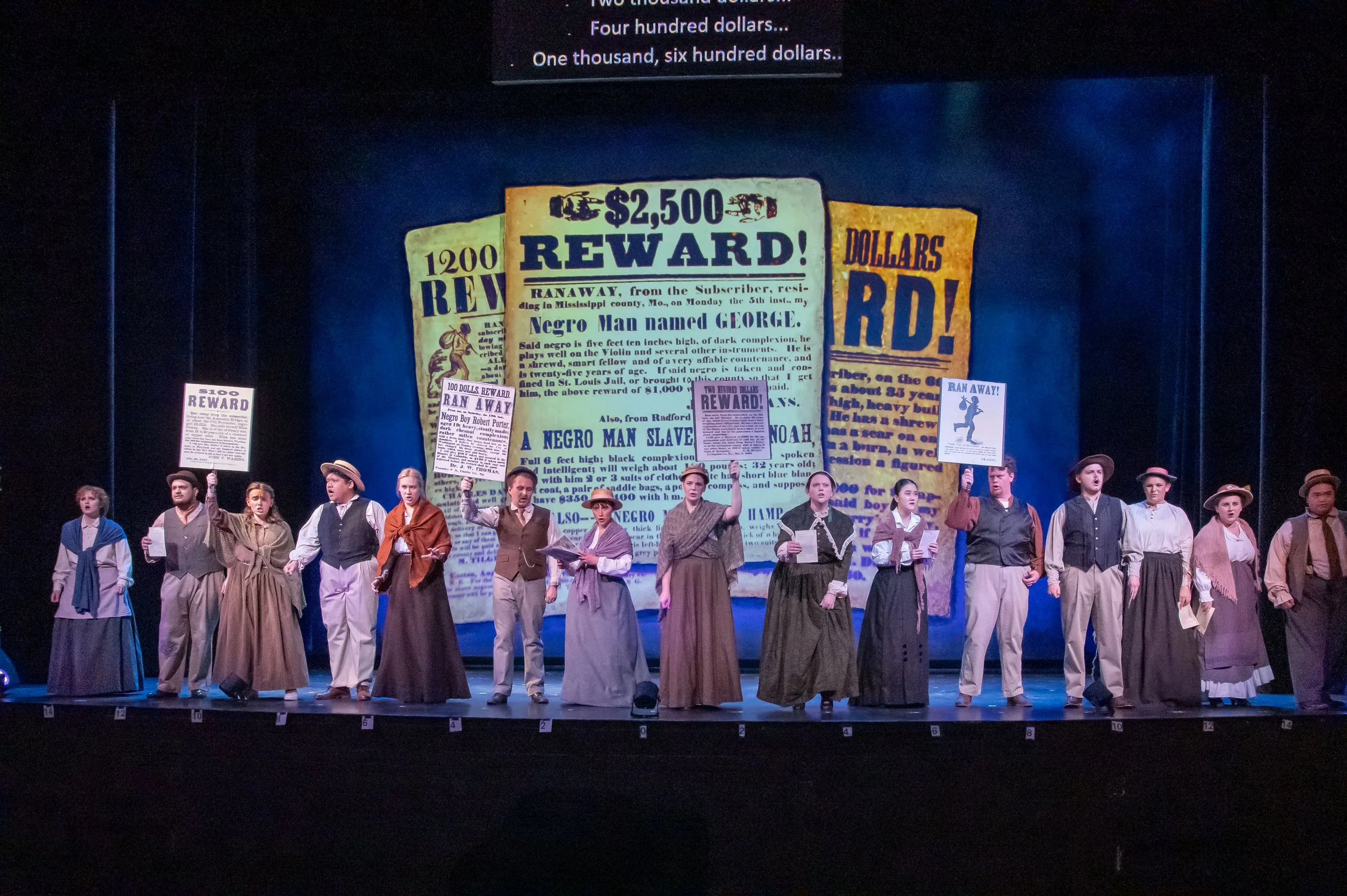 A theatrical stage with performers dressed in historical costumes. The background features a large screen displaying historic wanted posters. The performers are holding up smaller signs related to rewards and historical notices.