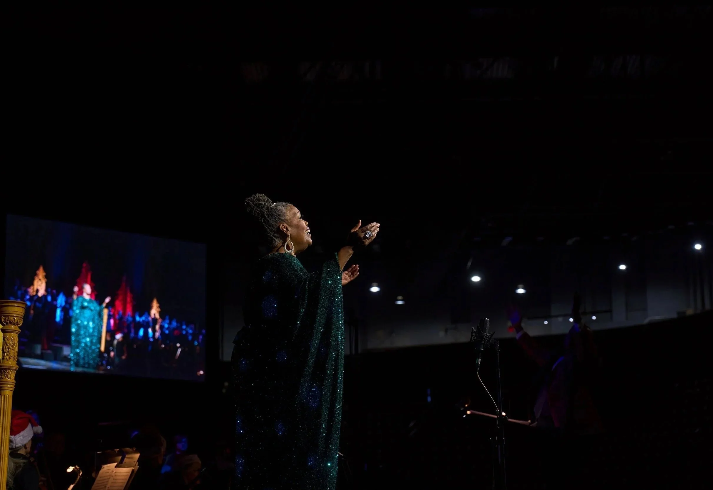 A woman singing on stage during a performance, wearing a sparkly green dress, with a choir or backup singers in the background in similar dresses, and Christmas decorations visible.