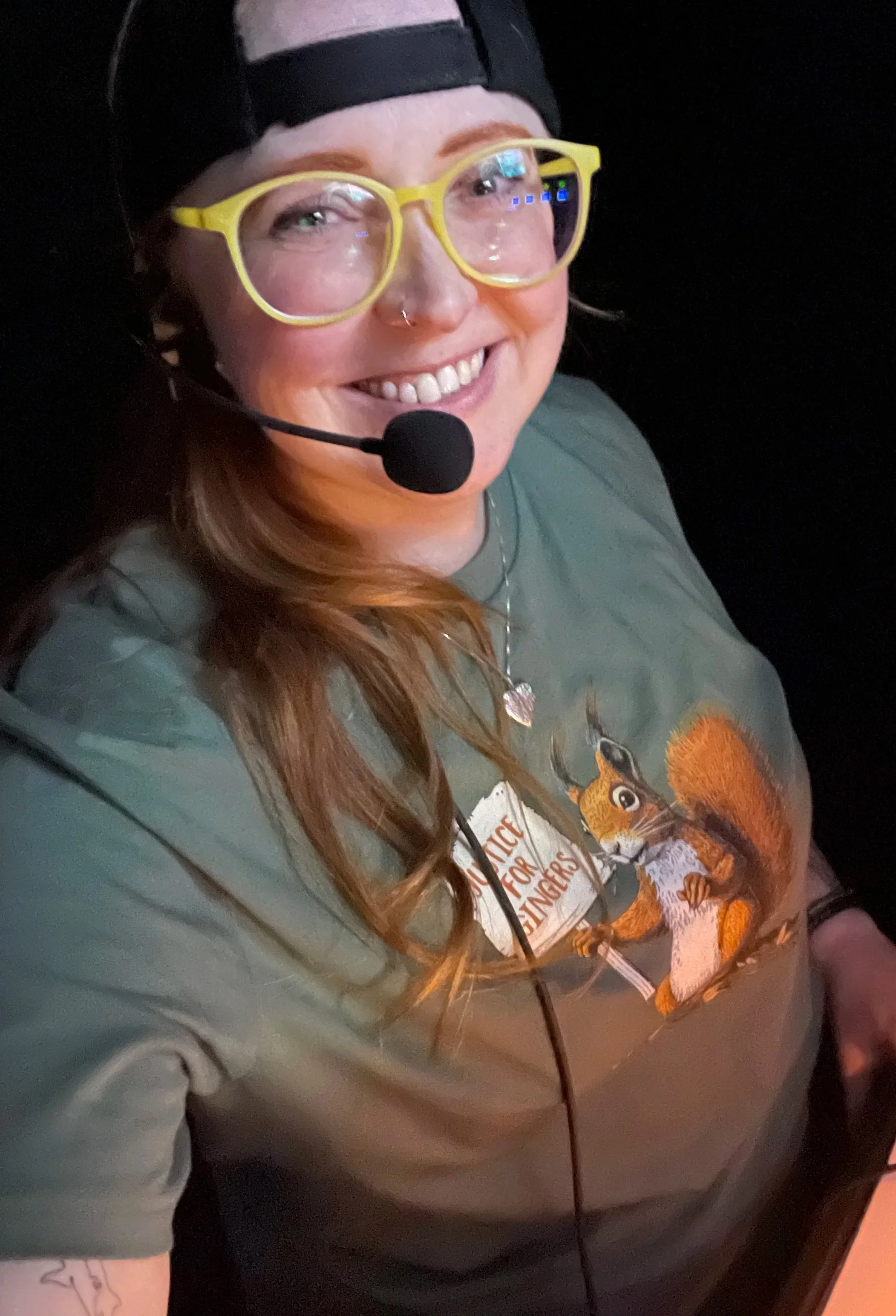 A woman with long, red hair, smiling, wearing bright yellow glasses, a black cap backwards, a gray T-shirt with a squirrel graphic, a necklace with a heart pendant, and a microphone headset.