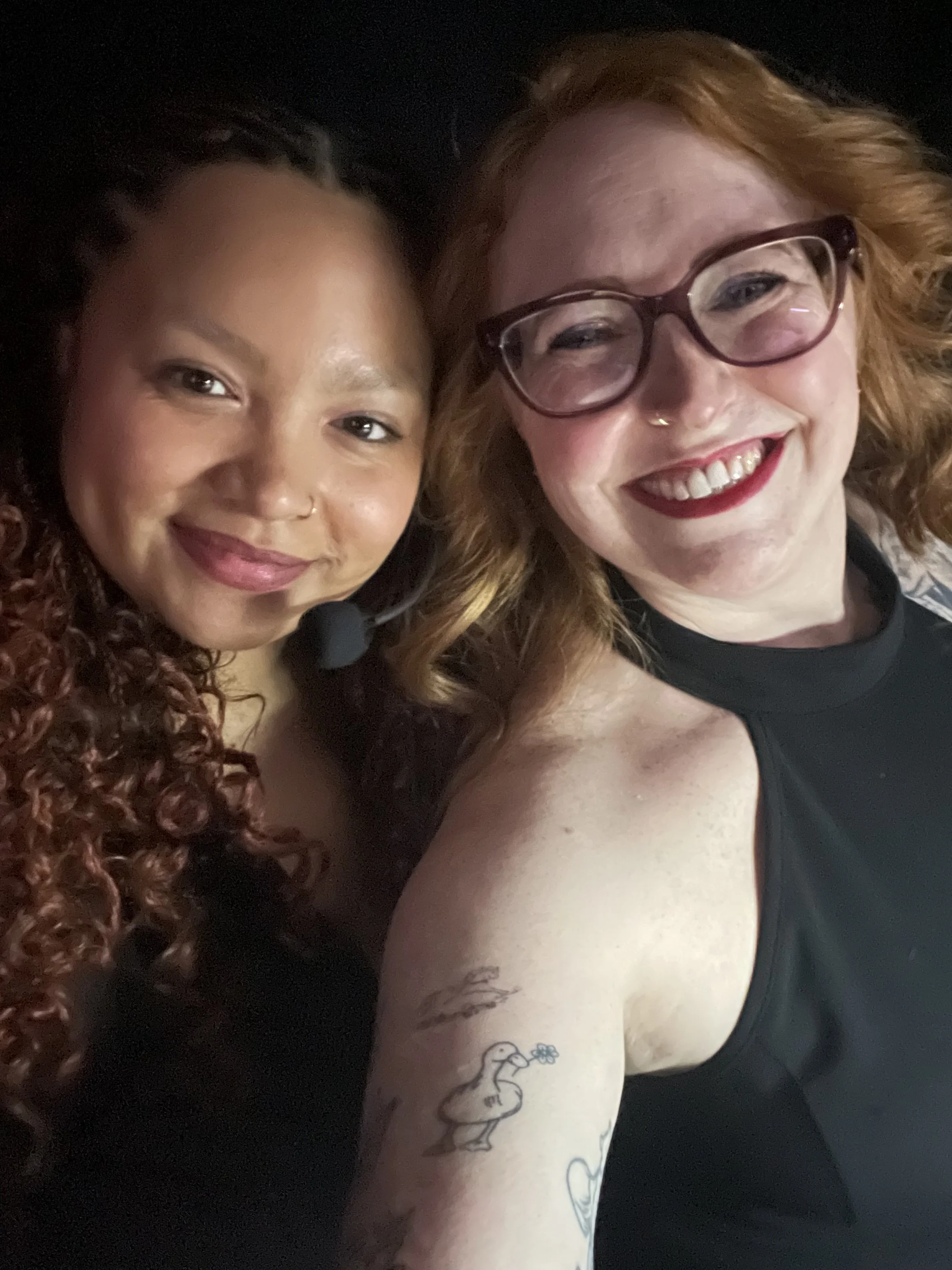 Close-up selfie of two smiling women with curly hair, one with dark skin and dark brown hair, the other with fair skin and red hair, wearing glasses, taken in a dimly lit setting.