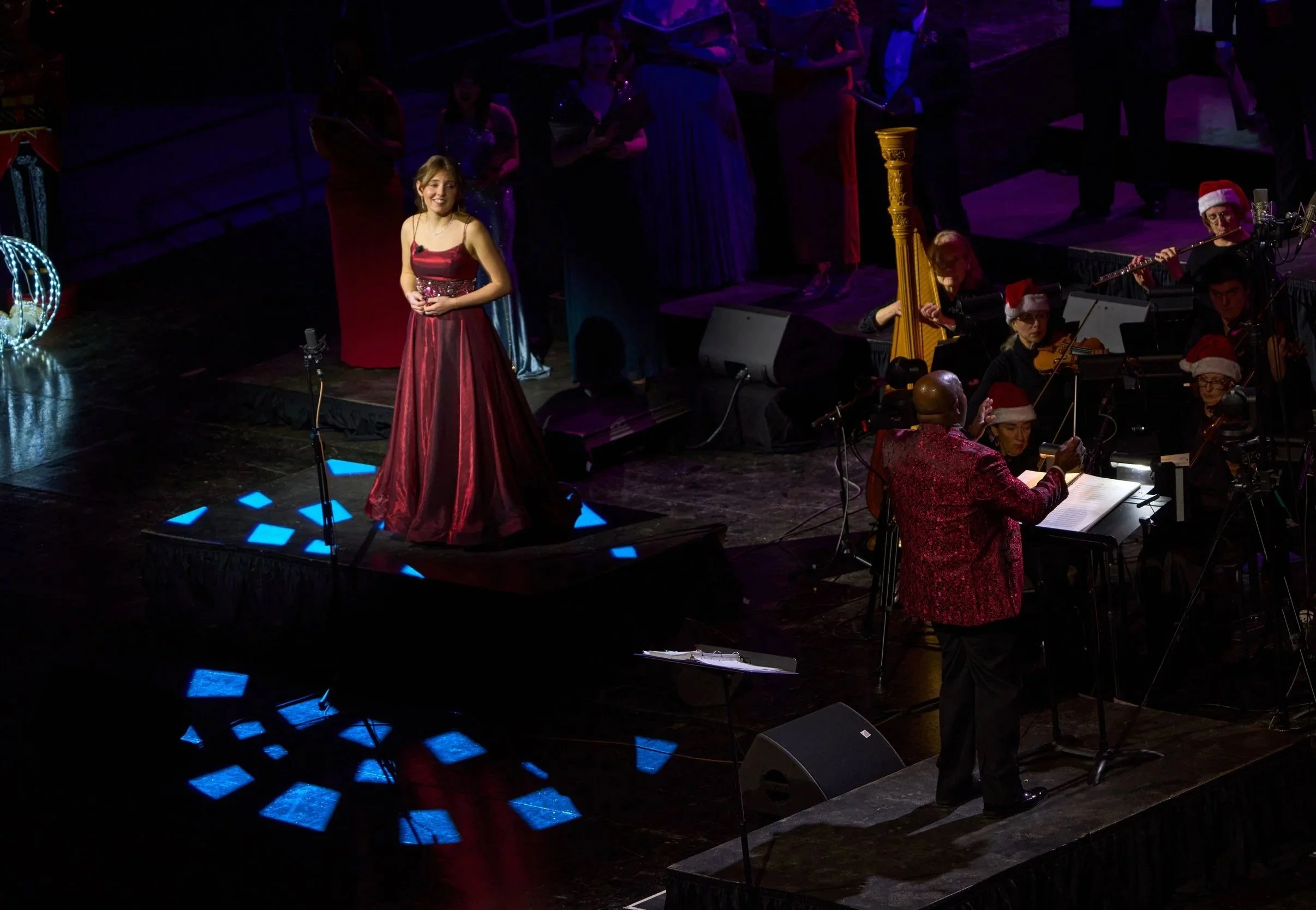 A woman in a red dress standing on a stage with a choir and musicians, some wearing Santa hats, during a holiday concert. Christmas decorations, including a lit ornament, are visible on the stage.