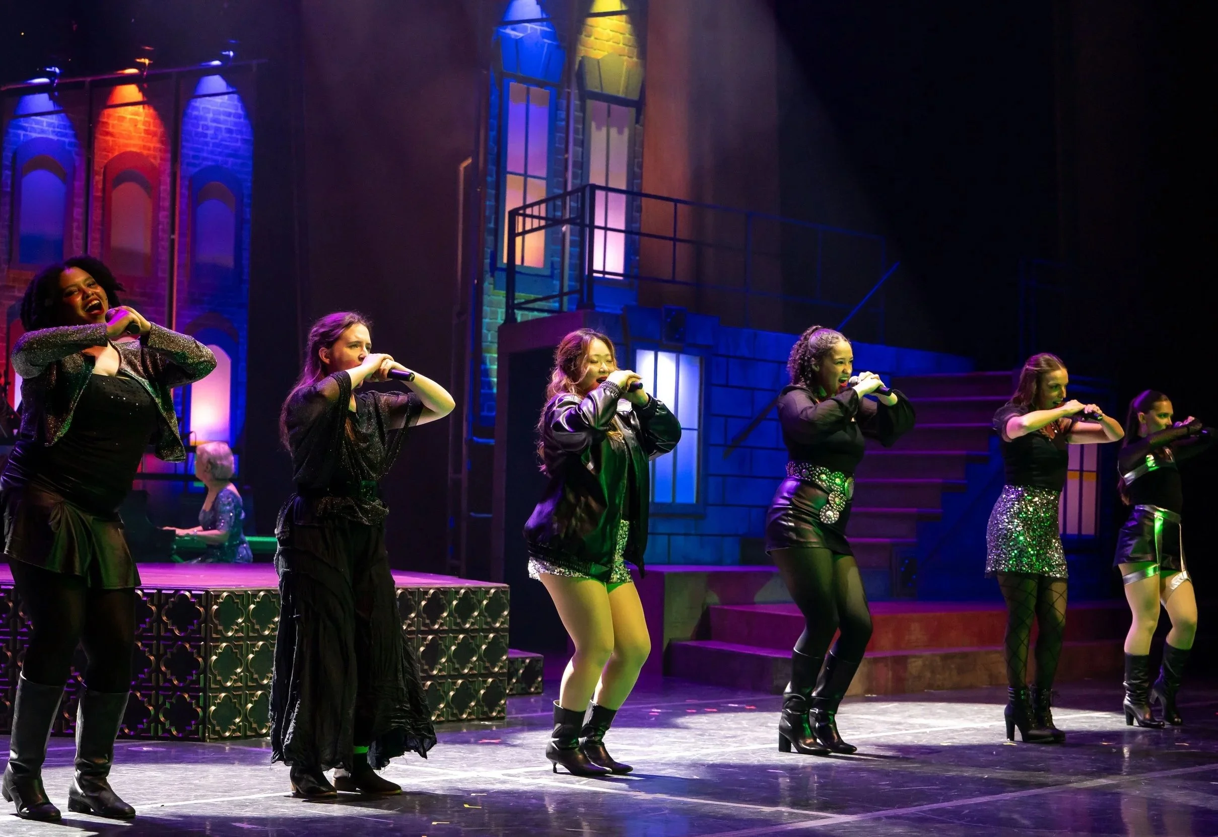 A group of women performing a dance routine on stage. They are wearing black outfits with boots and sequined accessories. The background features colorful lighting and a brick wall with large windows.