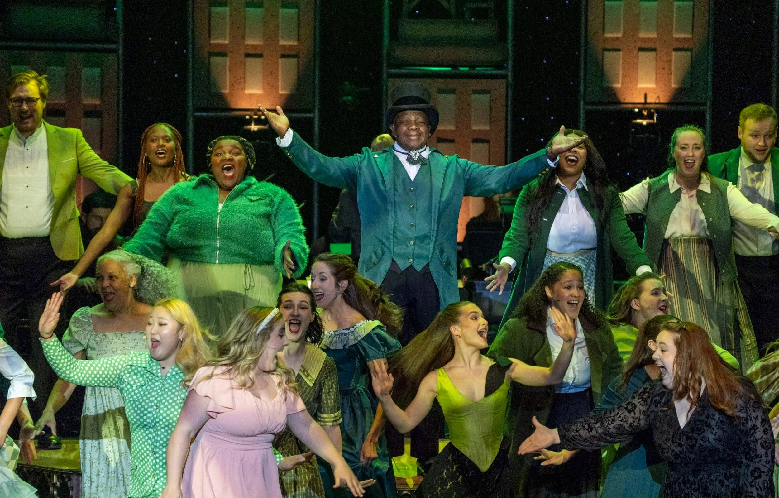 A diverse group of performers on stage, smiling and dancing in colorful costumes during a theatrical production.