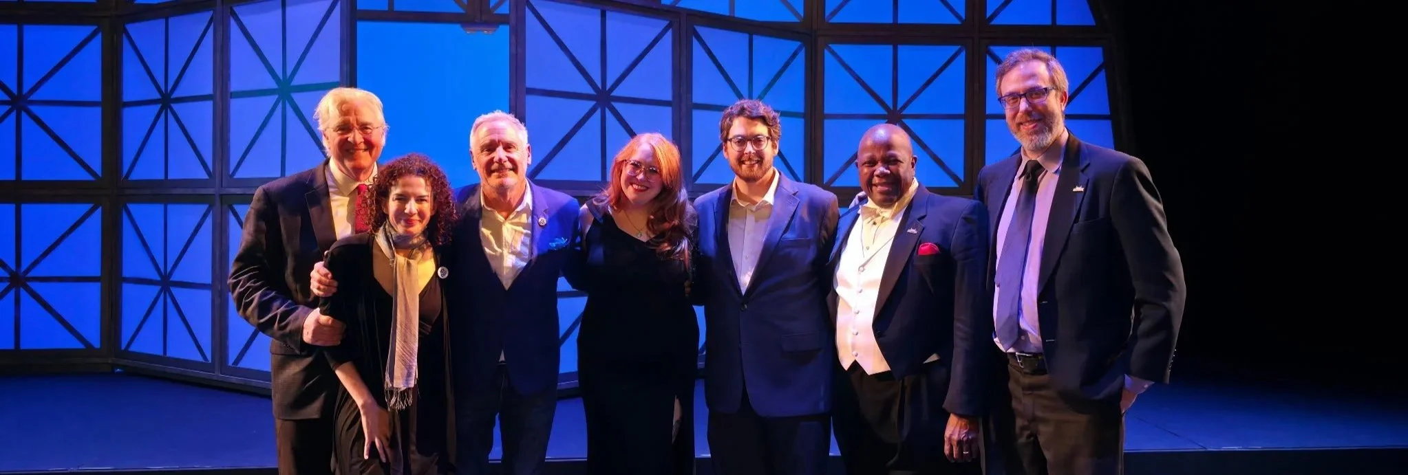 Group of seven people in formal attire standing together on a stage with blue geometric background.