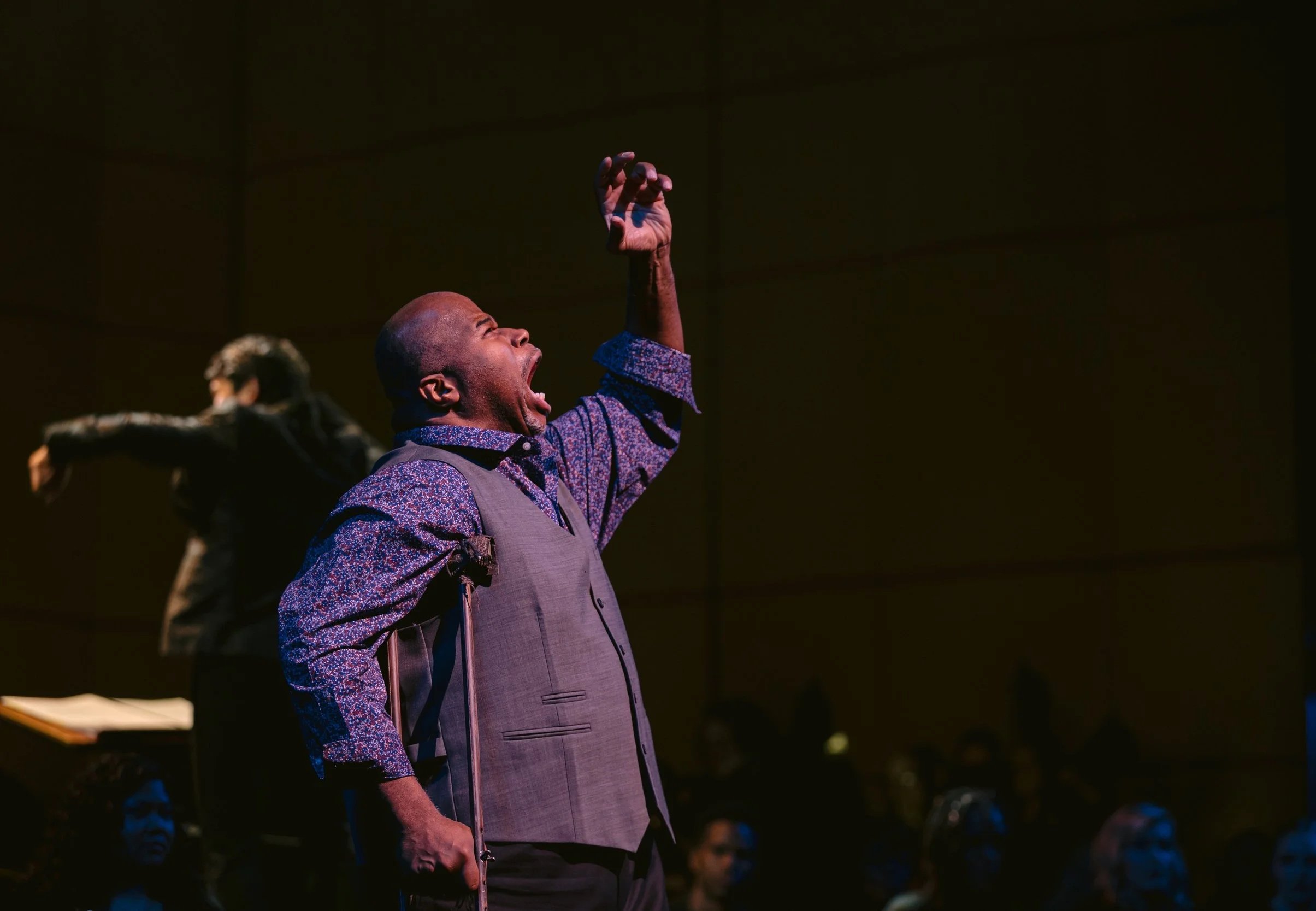 A man with a gray vest passionately singing on stage, with one arm raised and mouth open in front of a dark background and audience.