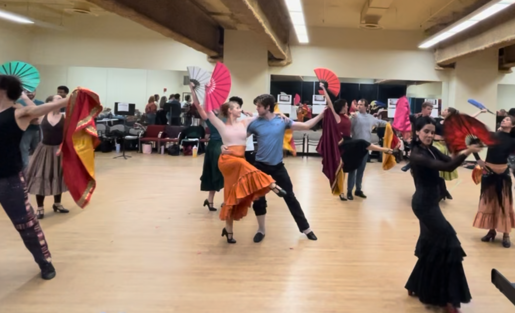 Couples dancing flamenco in a dance studio, holding colorful fans, with some dancers wearing traditional flamenco dresses.