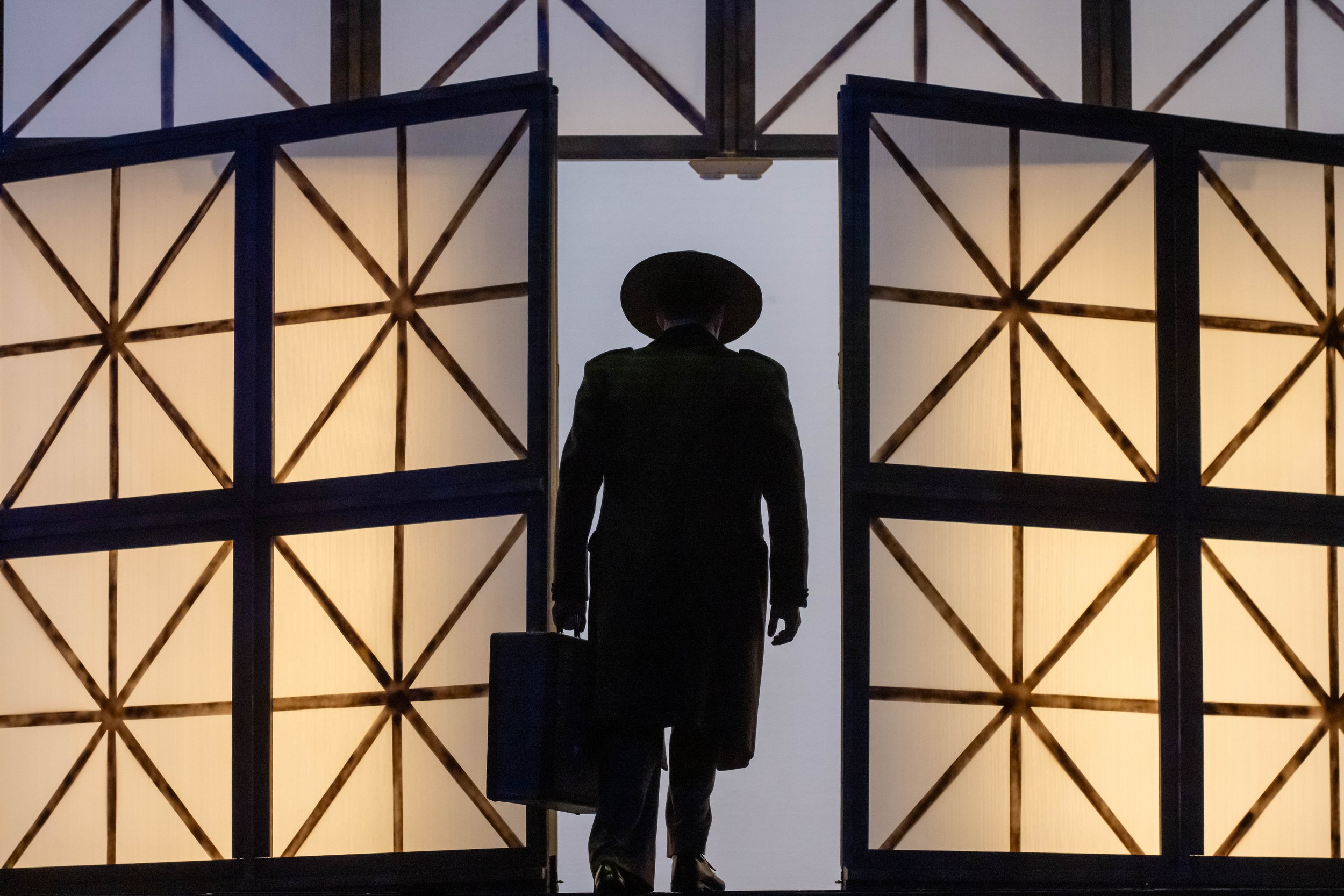 A person dressed in black with a wide-brimmed hat standing behind a partially open doorway or partition, with geometric lattice patterns and backlit panels creating a silhouette effect.