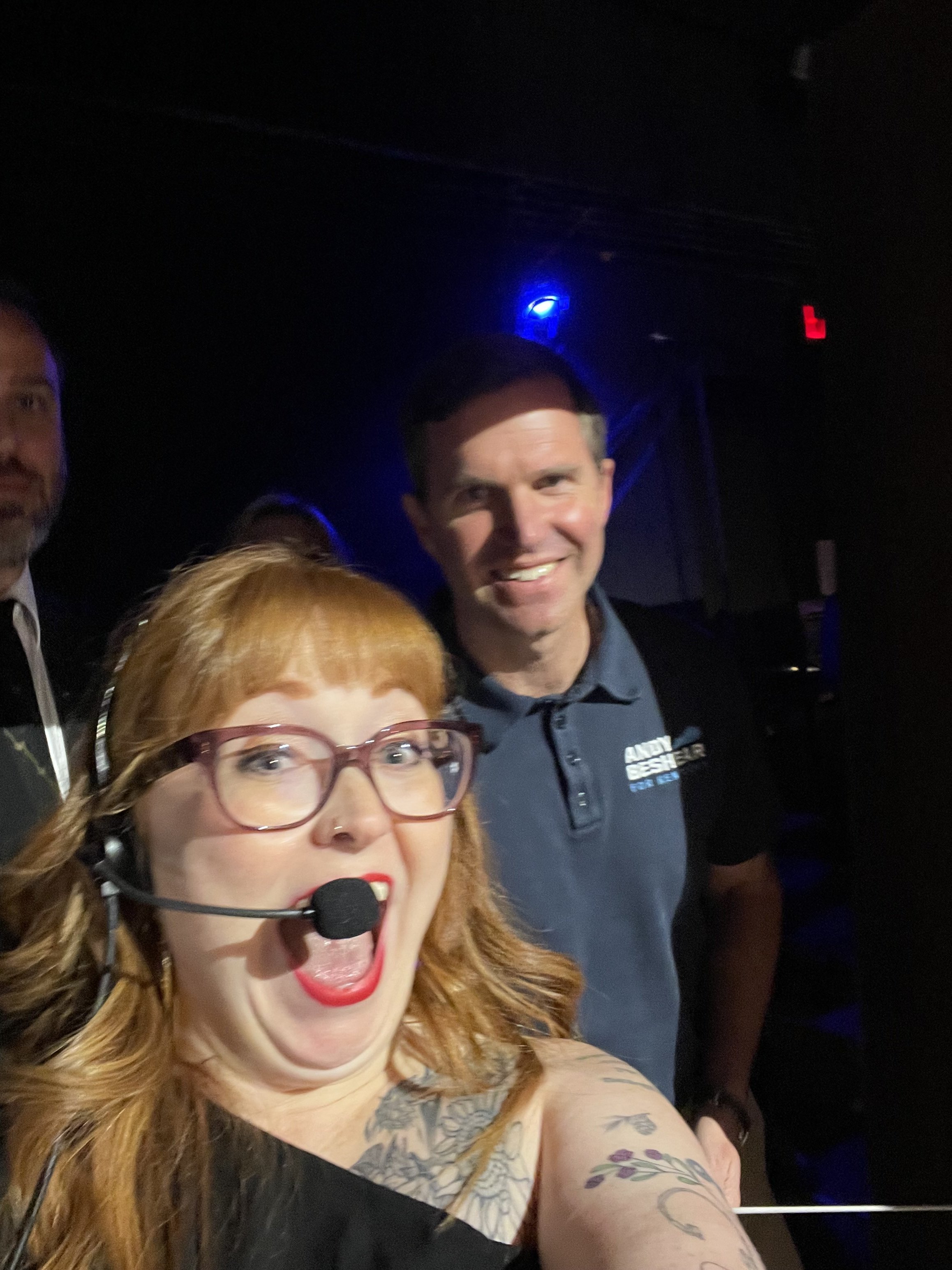 A woman with red hair, glasses, and tattoos on her arm taking a selfie with two smiling men in a dark indoor setting with blue and red lights.