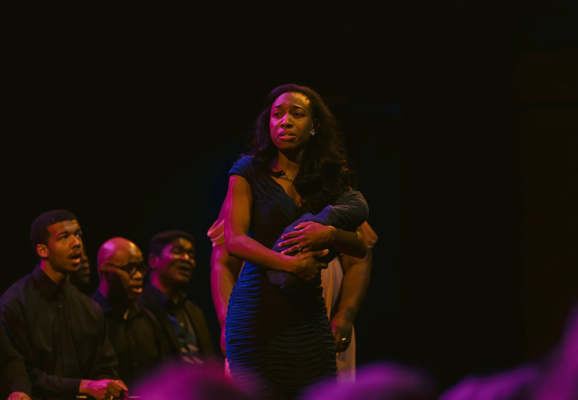 A woman with long curly hair wearing a blue dress onstage holding a doll, appearing pensive. Several people are seated around her, some looking toward her, in a dark setting with stage lighting.
