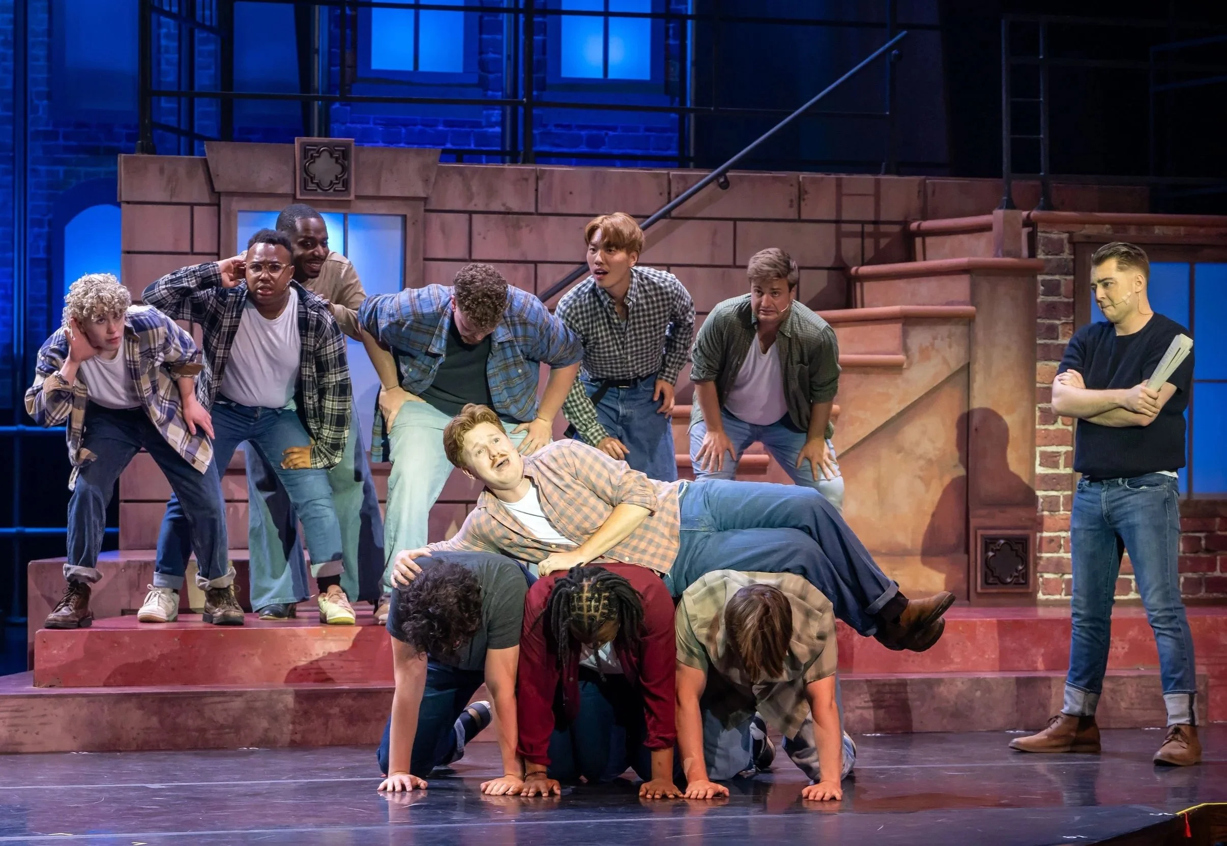 A group of performers on stage with stairs and brick walls. Some are on the stairs, others are on the floor, and a few are in mid-action.