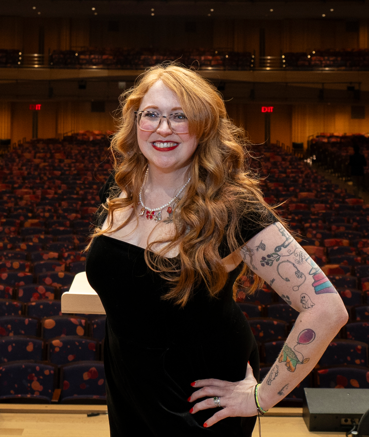 A woman with long red hair, glasses, and tattoos on her arms, smiling in an auditorium with empty seats in the background.