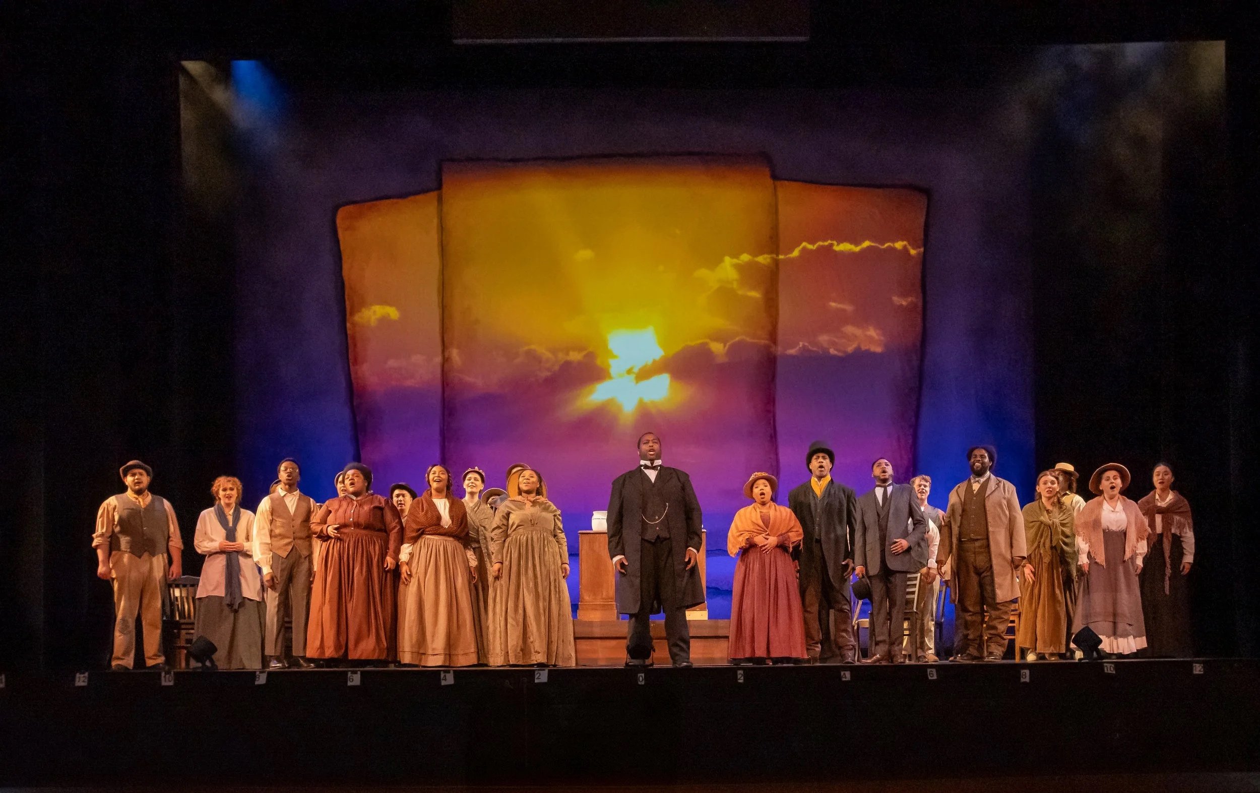 A group of diverse people performing on stage dressed in 19th-century clothing, with a backdrop depicting a sunset or sunrise.