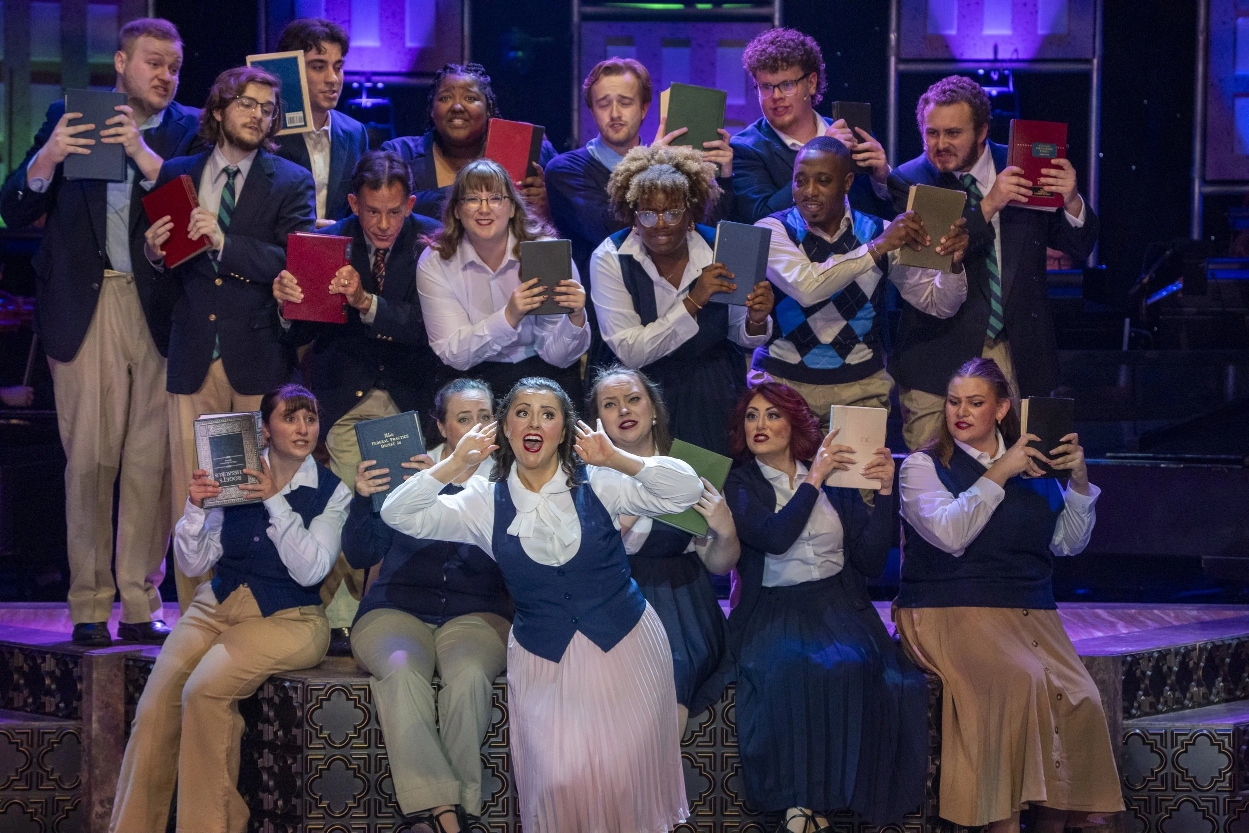 Group of actors on stage in performance, some holding books, displaying various facial expressions and gestures.