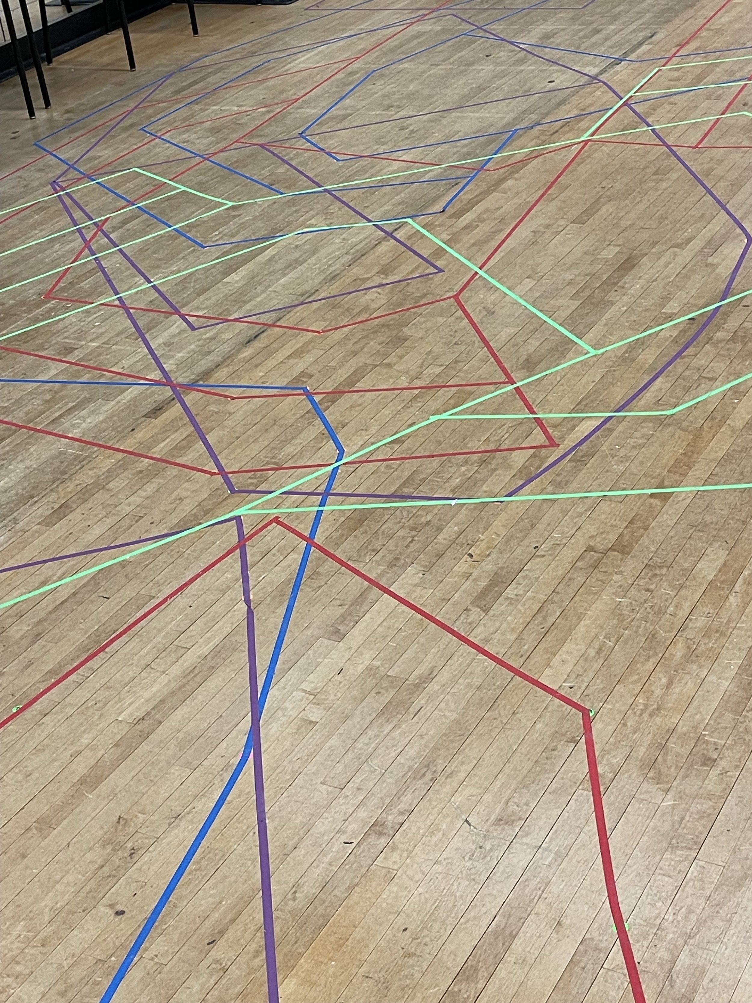 Colorful glow-in-the-dark tape forming an abstract geometric pattern on a wooden floor, with chairs and tables in the background.
