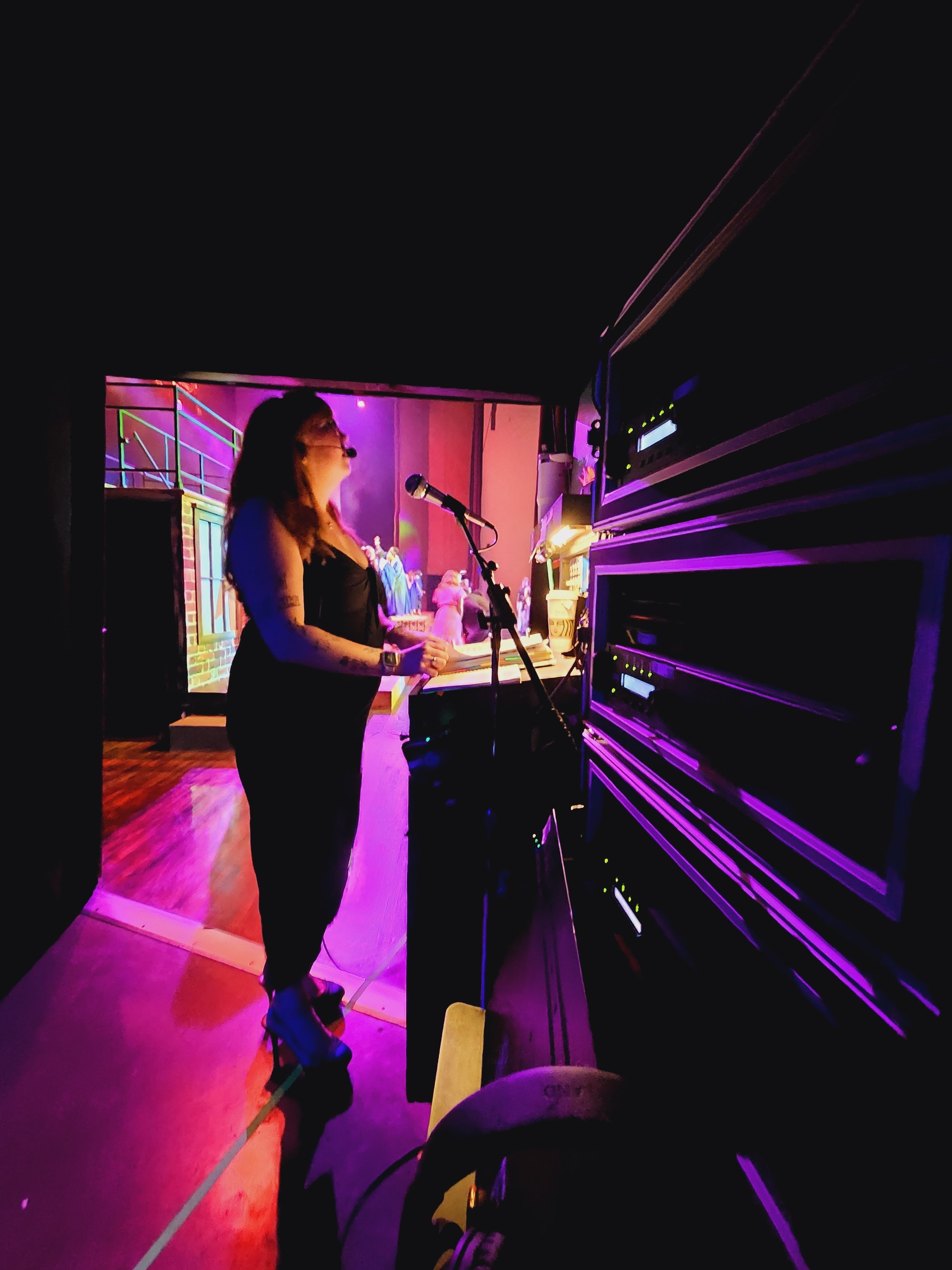 A woman in a black dress and glasses standing by a piano on a stage with purple and pink lighting, facing a microphone.