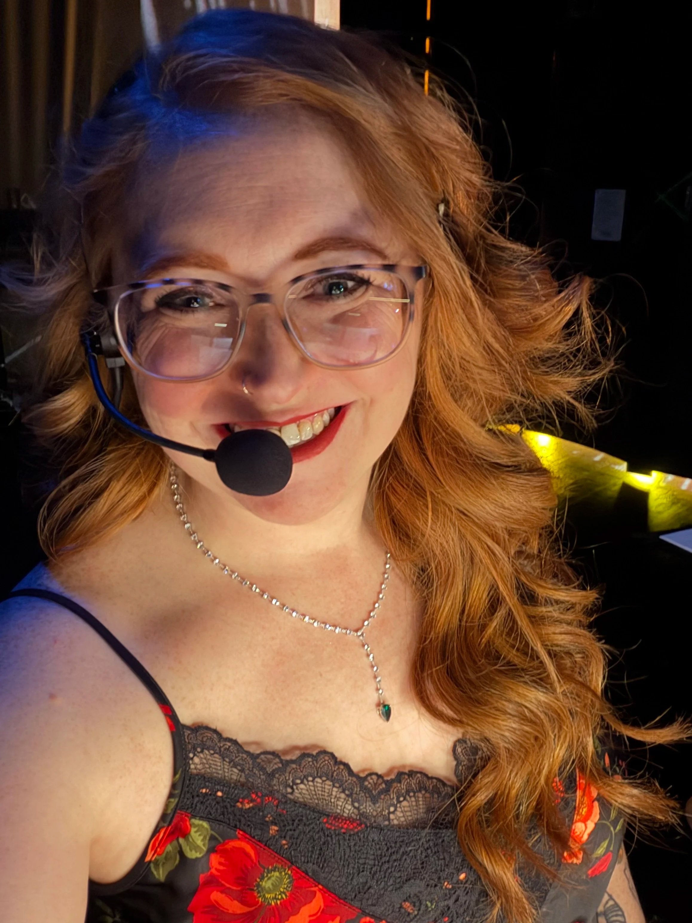 Close-up photo of a woman with red hair, wearing glasses, a microphone headset, and a black lace top with red and green floral print. She is smiling and looking into the camera.