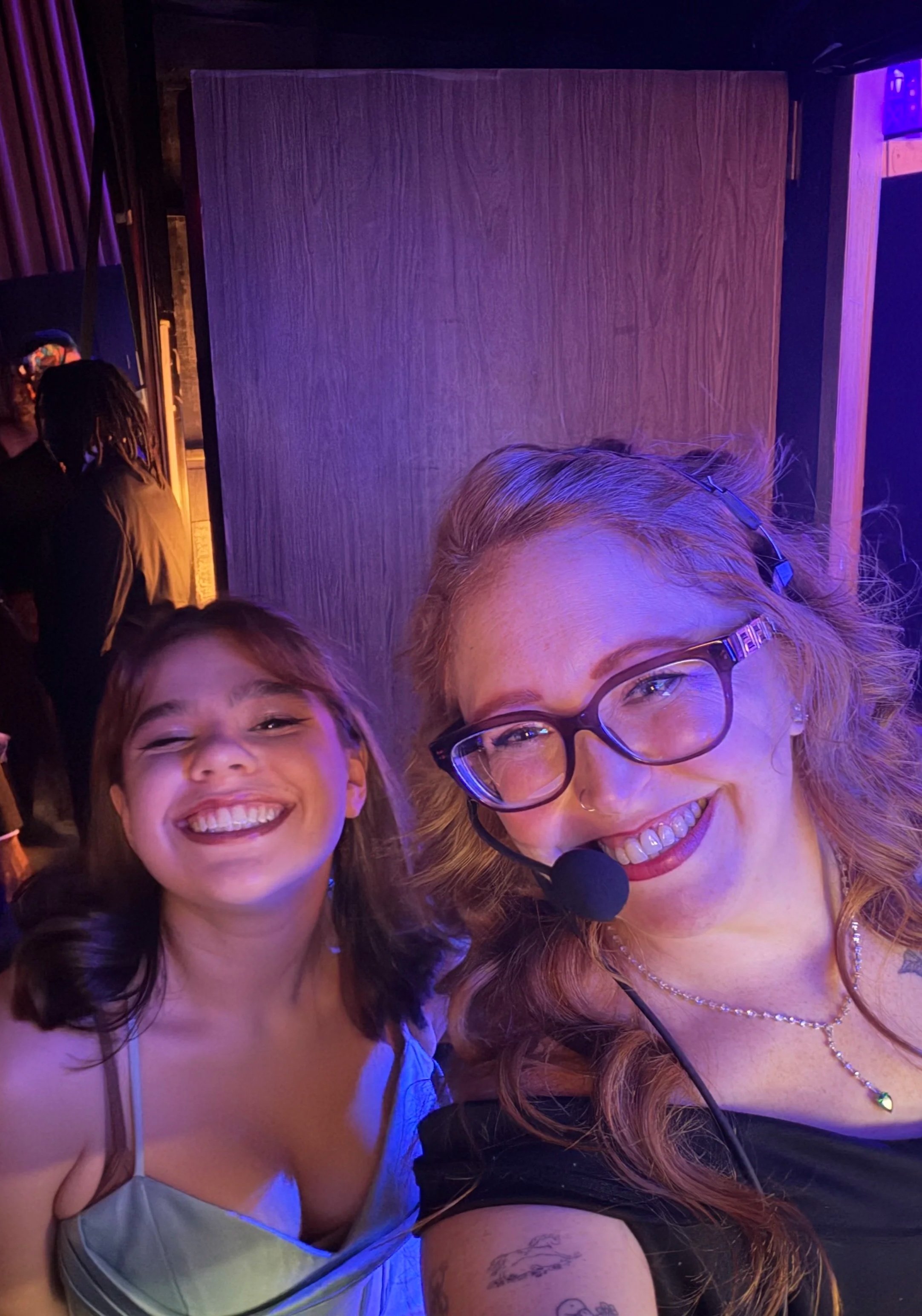 A happy young girl and a woman, both smiling at the camera, with the woman wearing glasses and a headset. The background shows dim lighting with purple accents.
