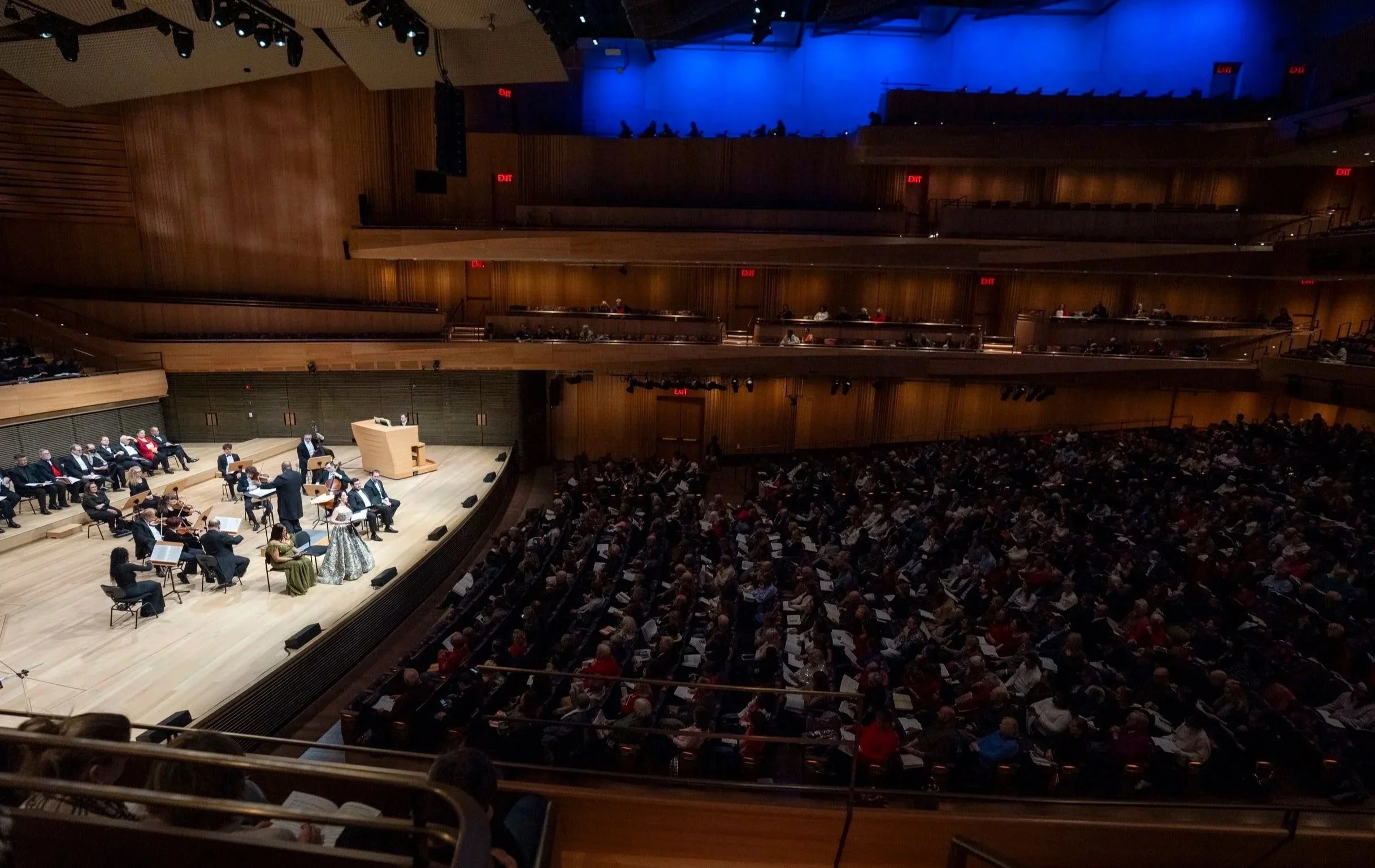 An auditorium with a stage where an orchestra is performing, and a conductor is leading them. The audience is seated and watching the performance, and the room has multiple levels with wooden paneling and emergency exit signs visible.
