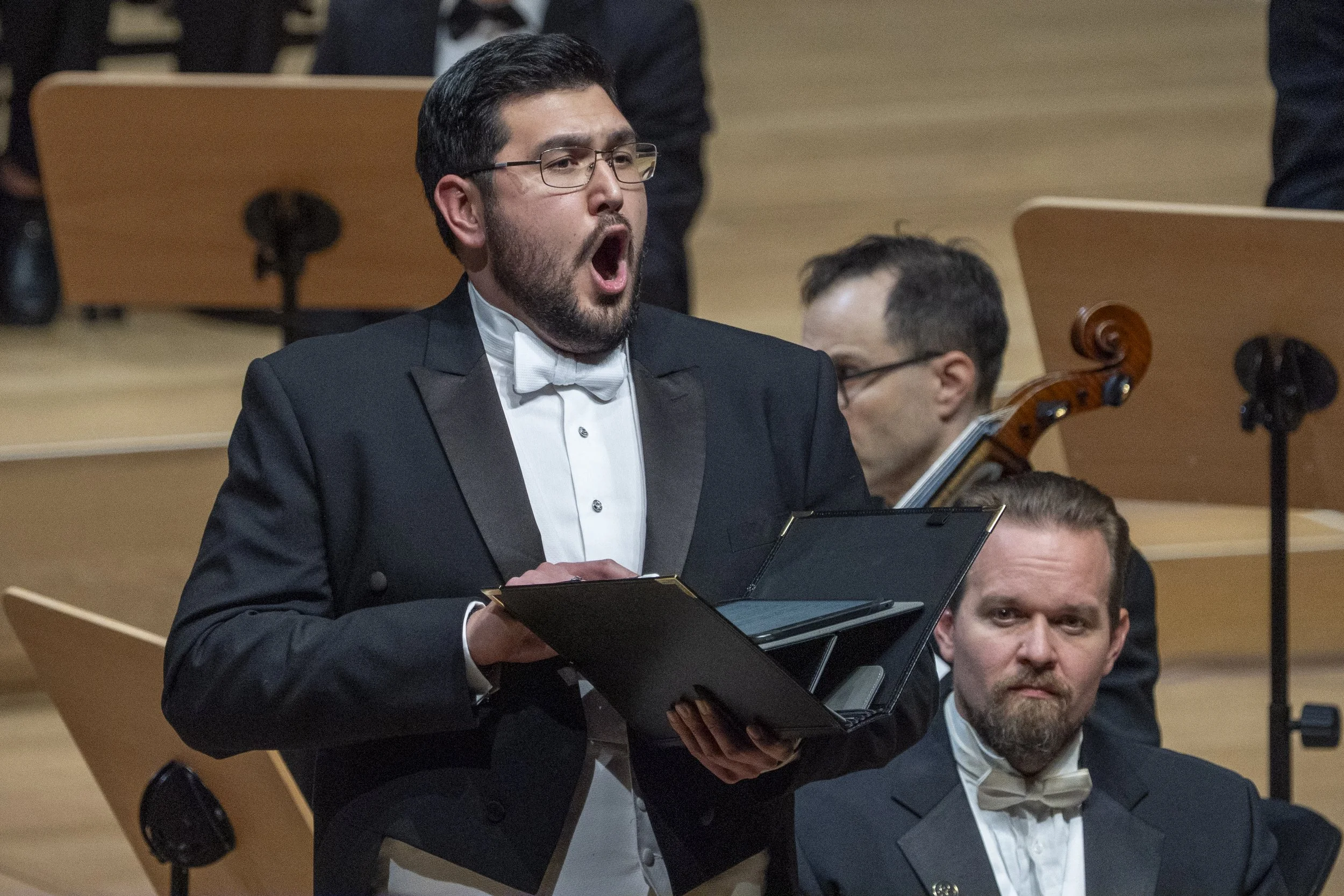 Man in formal attire, tuxedo and white bow tie, singing with an open mouth, holding music. He is wearing glasses and has dark hair and beard, seated among others in a concert hall with wooden seats.