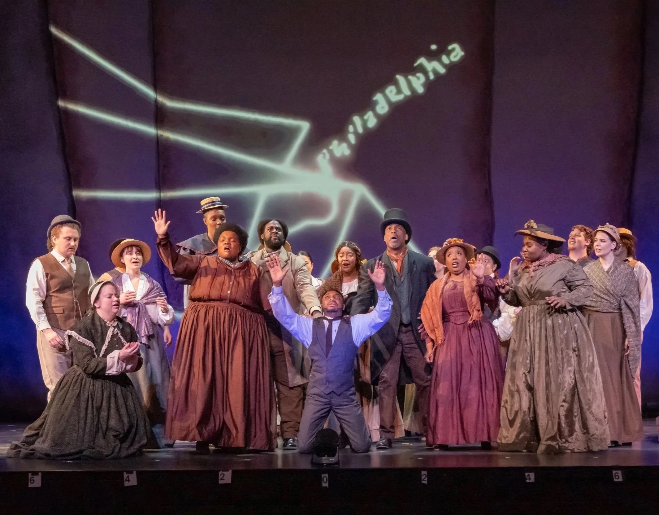 A group of people on stage dressed in historical clothing, possibly from the 19th century, with some raising their hands. There is a dark curtain backdrop and a projected image.