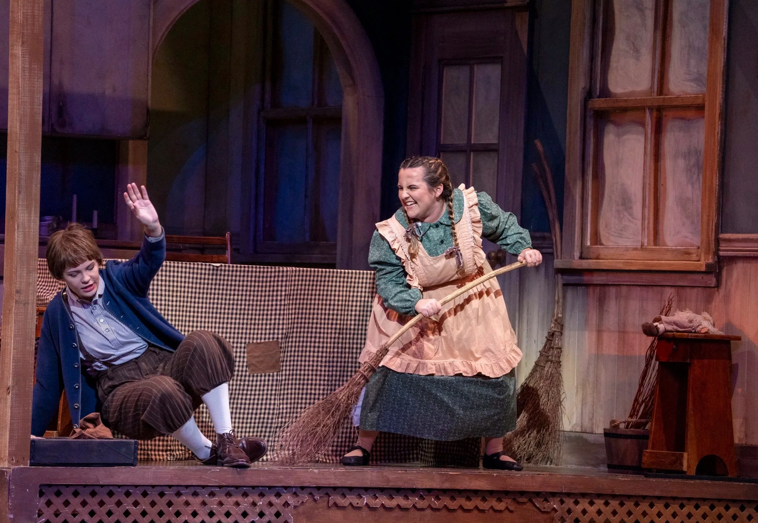 A scene featuring two performers on stage. One performer, a woman dressed in period costume with an apron, is holding a broom and smiling. The other actor, a young man in vintage clothing, is seated and raising his hand.