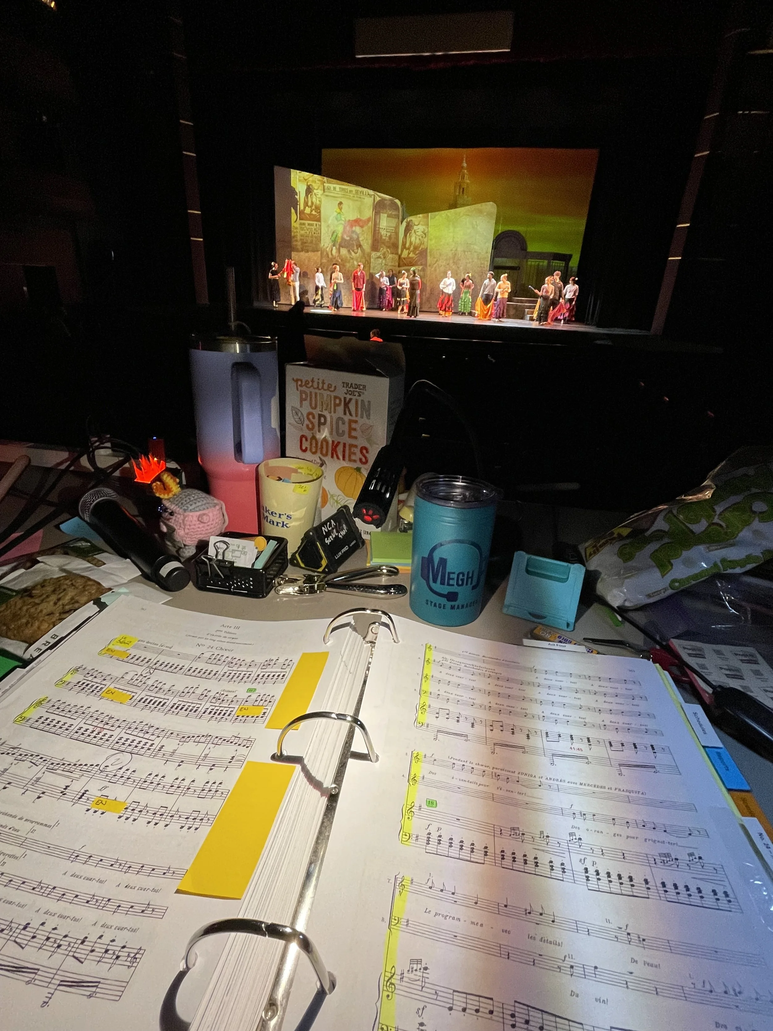 Open binder with sheet music and yellow highlighter on music pages, cluttered desk with oversized cup, snacks, keys, and miscellaneous items, stage with performers in colorful costumes performing a dance or theatrical scene in a theater setting.
