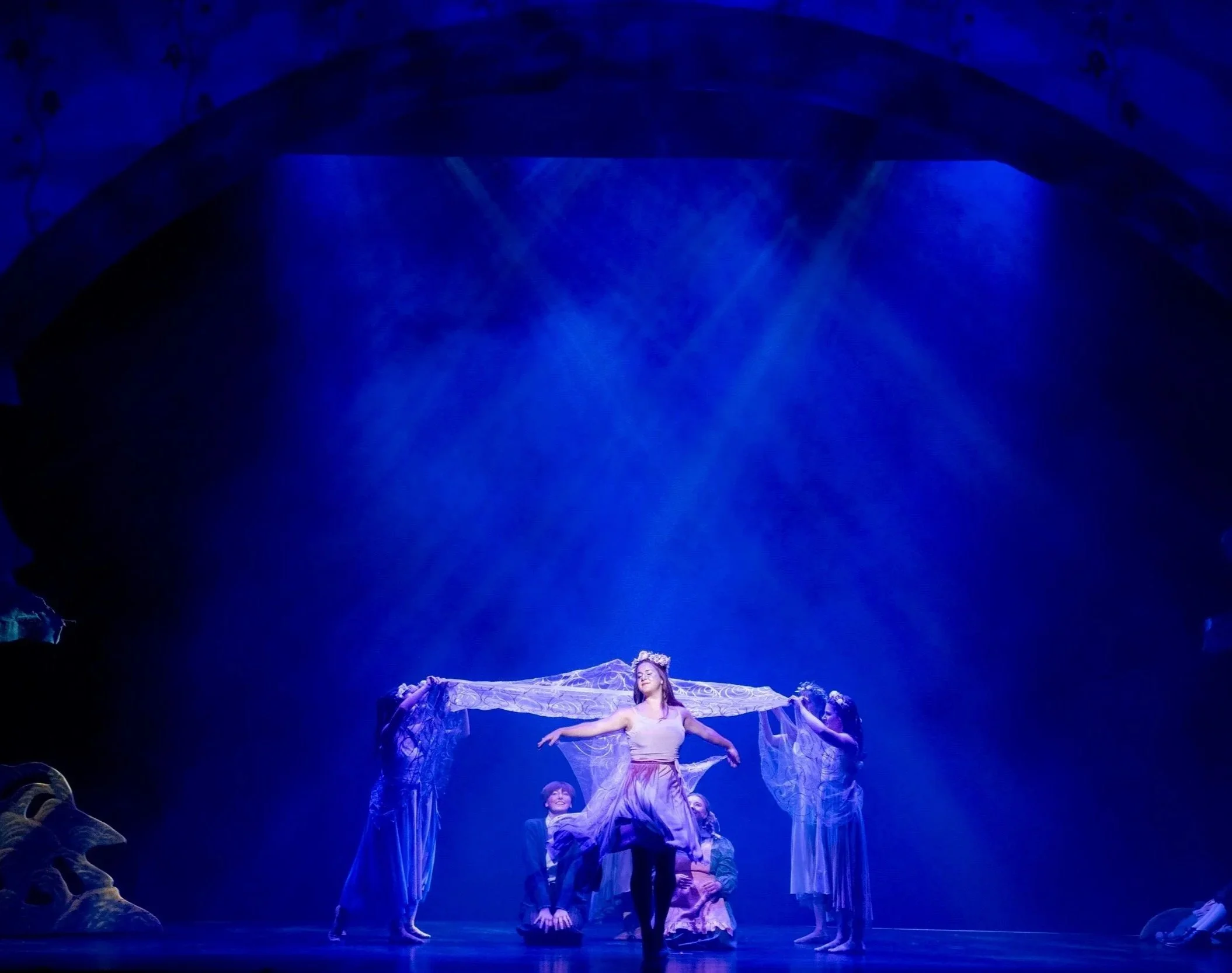 Performers dressed in flowing costumes on stage with blue lighting and a backdrop that resembles a starry night sky, during a theatrical performance.