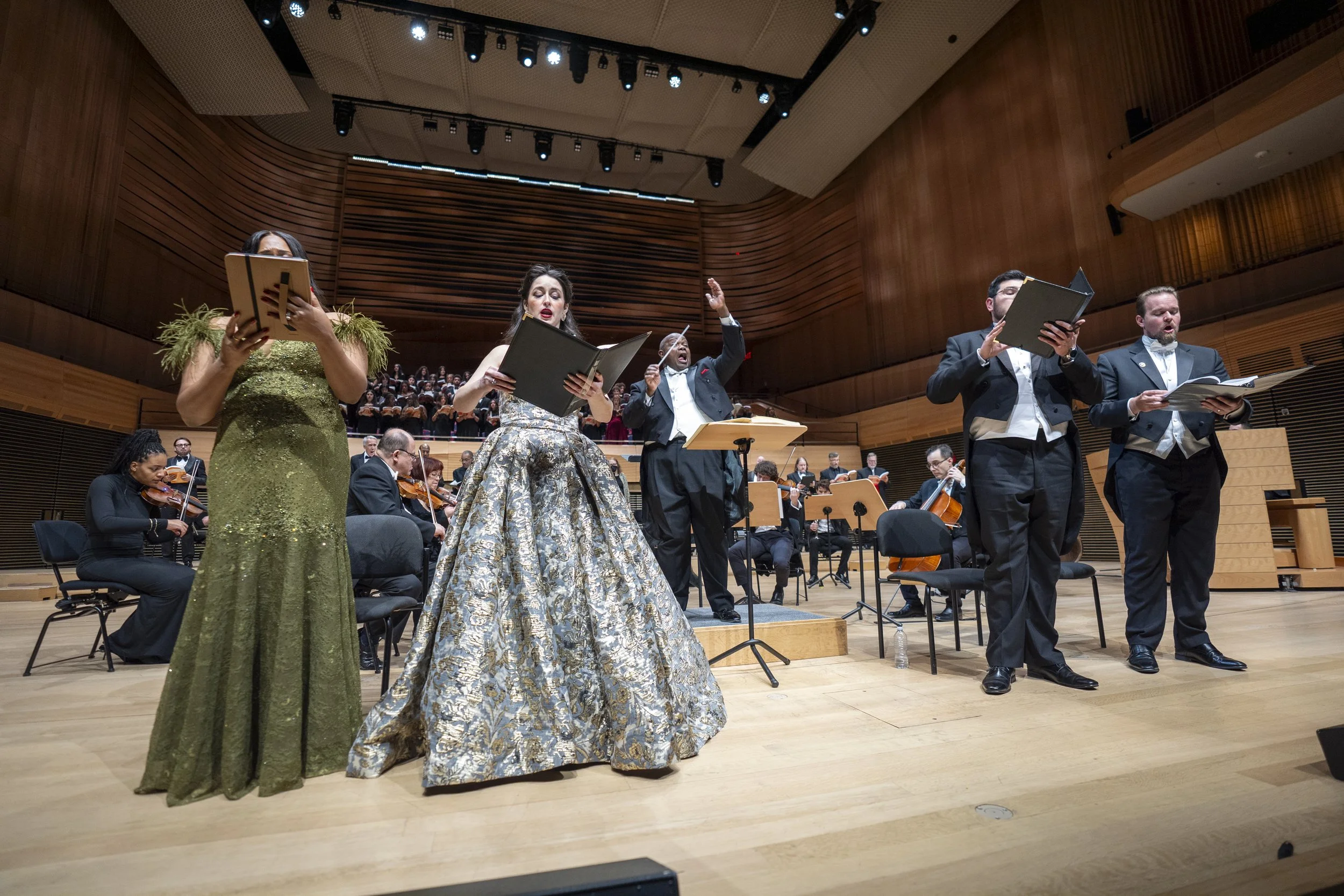 Orchestra performers on stage in concert hall, women in elegant dresses and men in tuxedos, with musicians playing string instruments in the background.