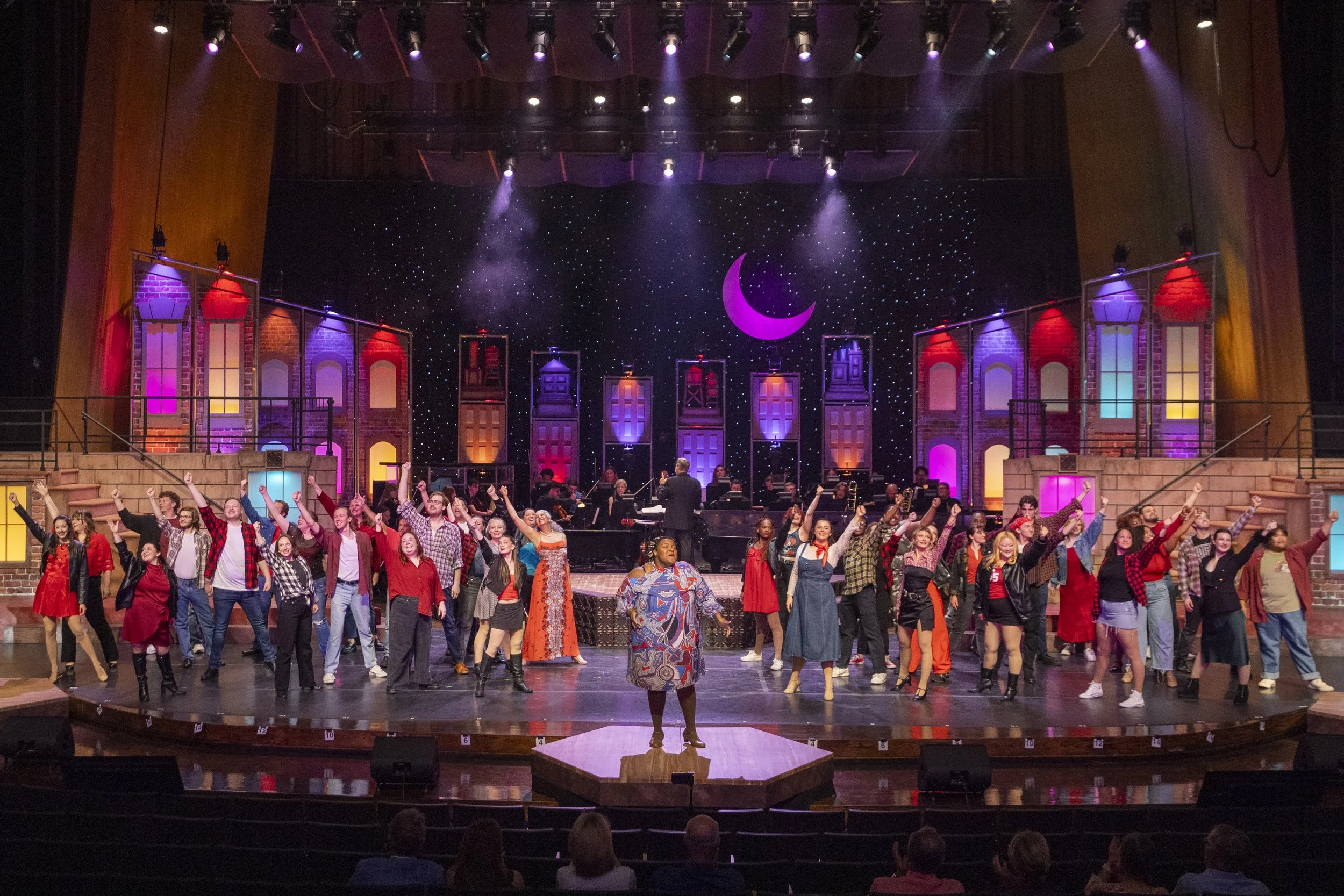 A stage in a theater with a large group of diverse performers, some dancing and some standing, under colorful lights. The stage backdrop features a crescent moon and starry night design, with colorful stained glass-style windows on the sides. Audience members are visible in the foreground.