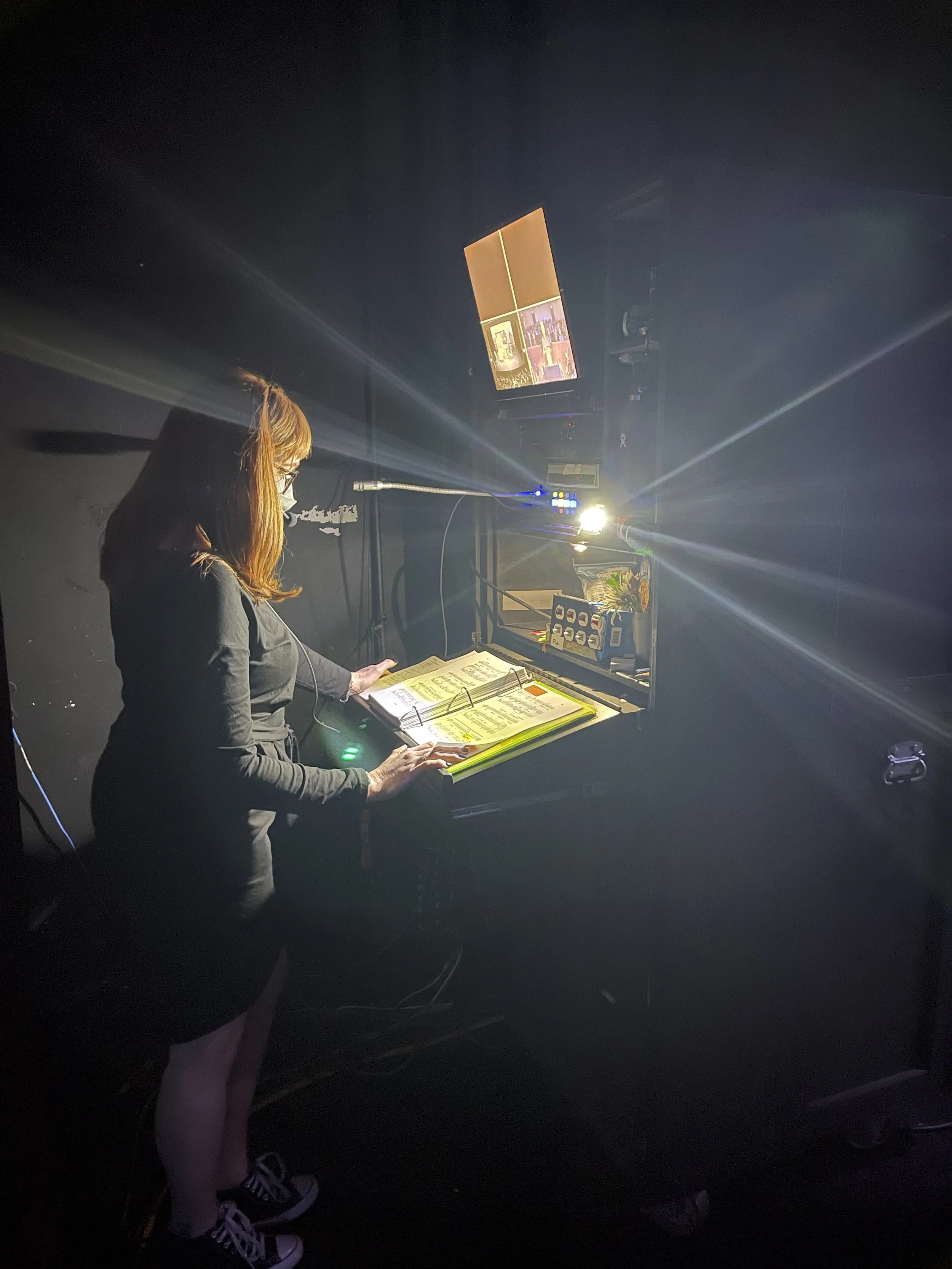 A woman wearing a face mask and glasses at a SM console backstage calling an operatic production