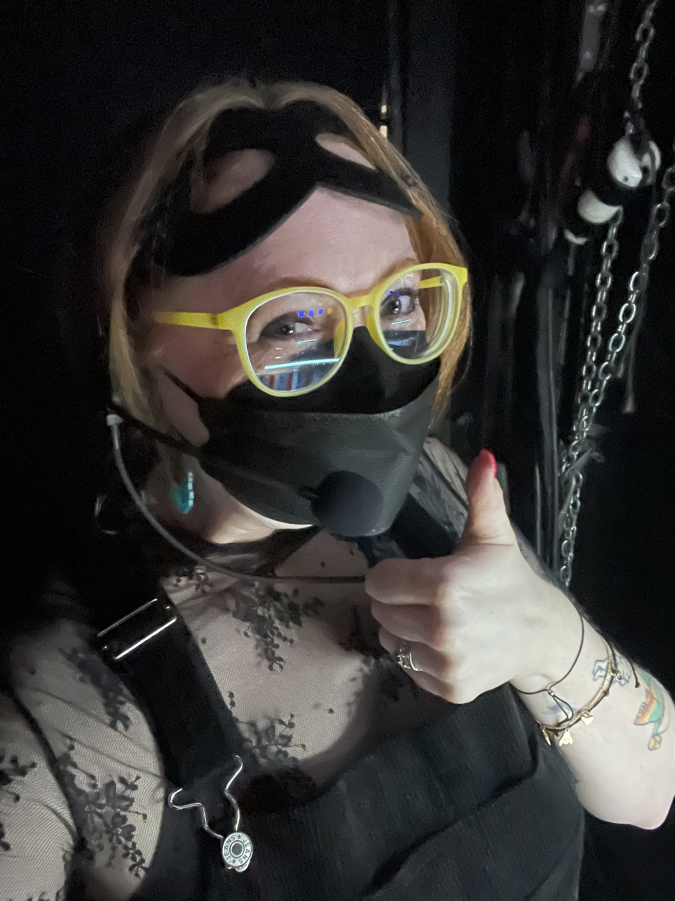 A woman with red hair, wearing yellow glasses, a black face mask, and a black lace top and giving a thumbs-up while backstage..