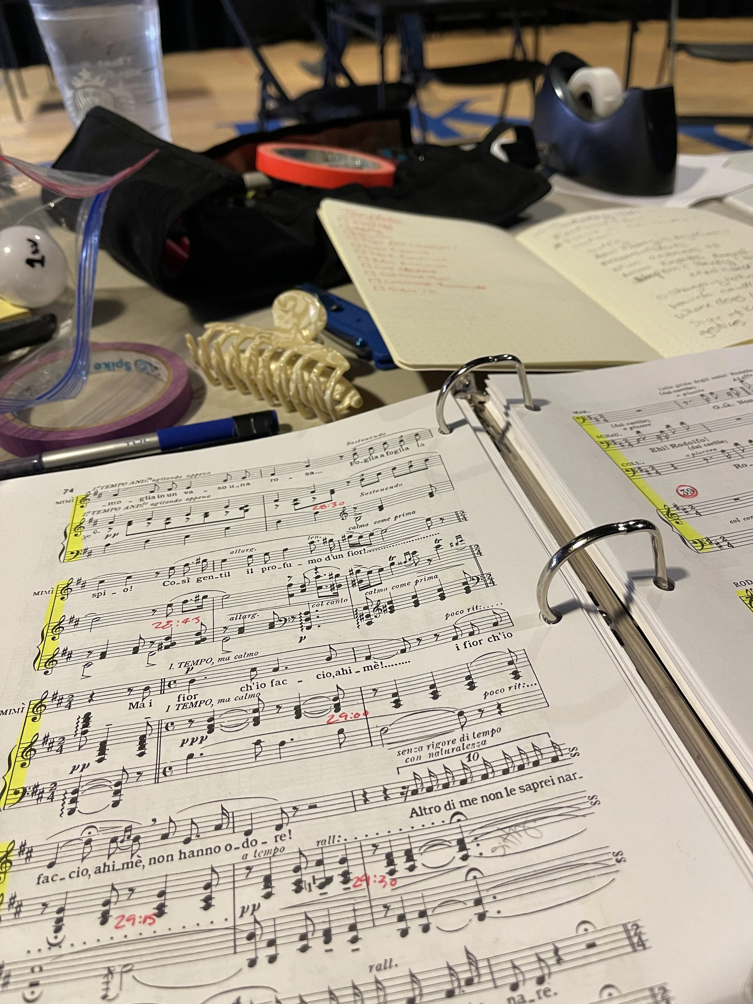 Open music sheet with notes and annotations, surrounded by miscellaneous items including a blue pen and a gold skeleton-shaped object on a cluttered desk.