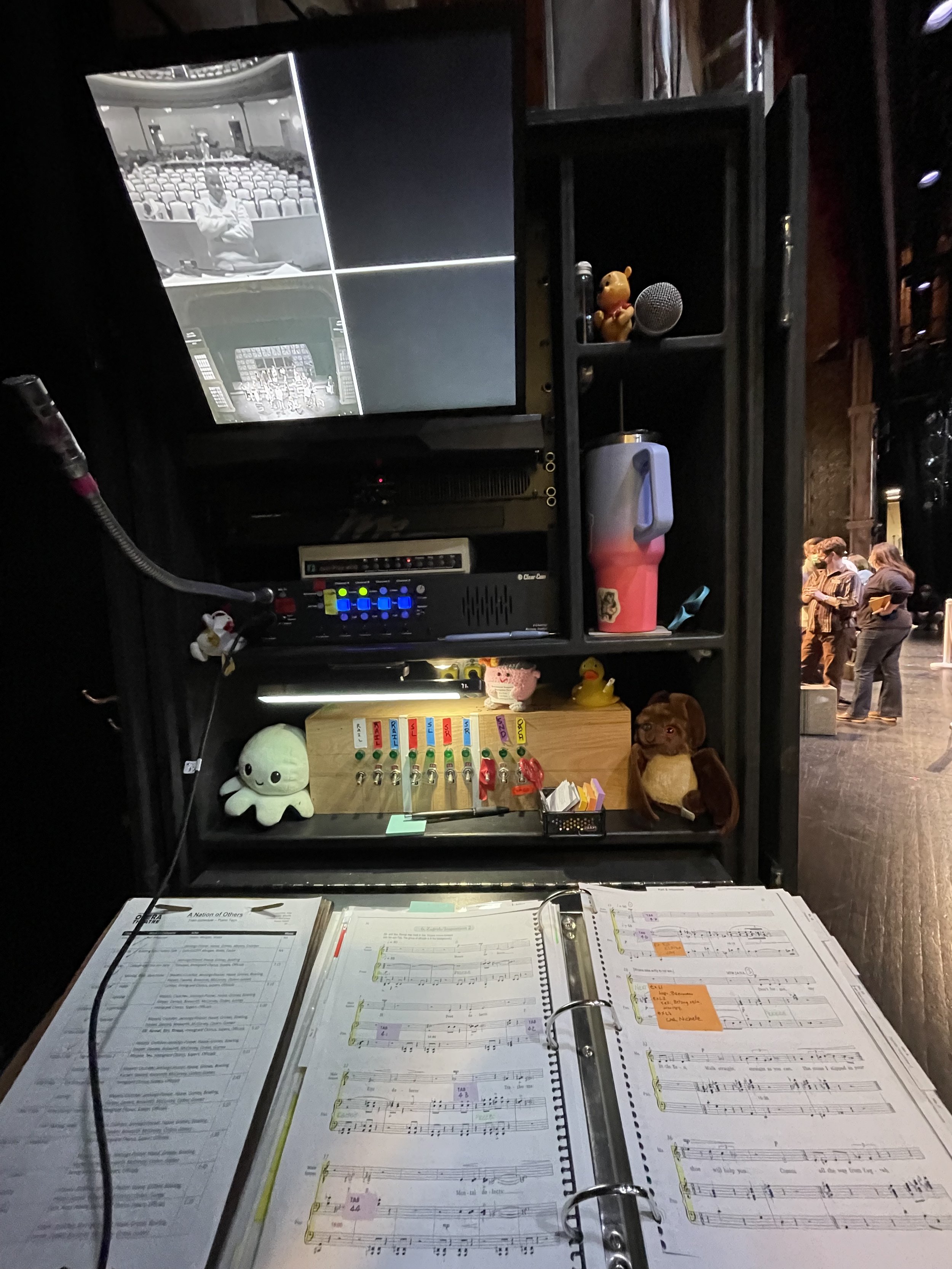A close-up of sheet music, two plush toys, and a gathering of people in the background.