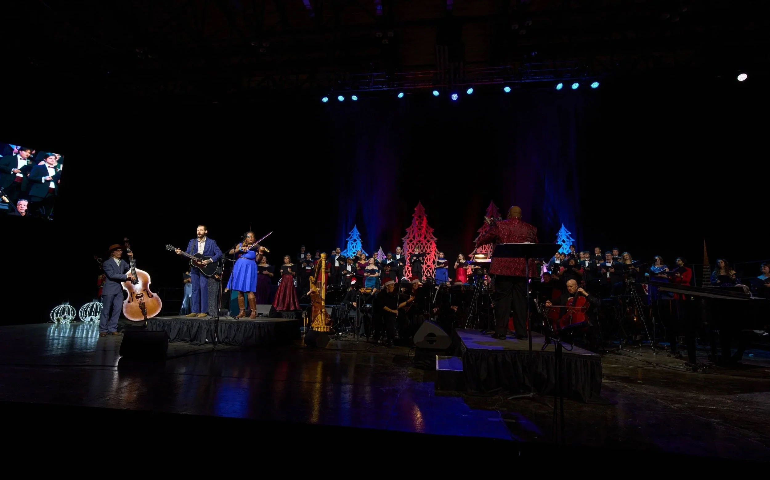 Large choir and orchestra performing on stage decorated with holiday-themed ornaments and trees, under blue and red lighting, during a concert.