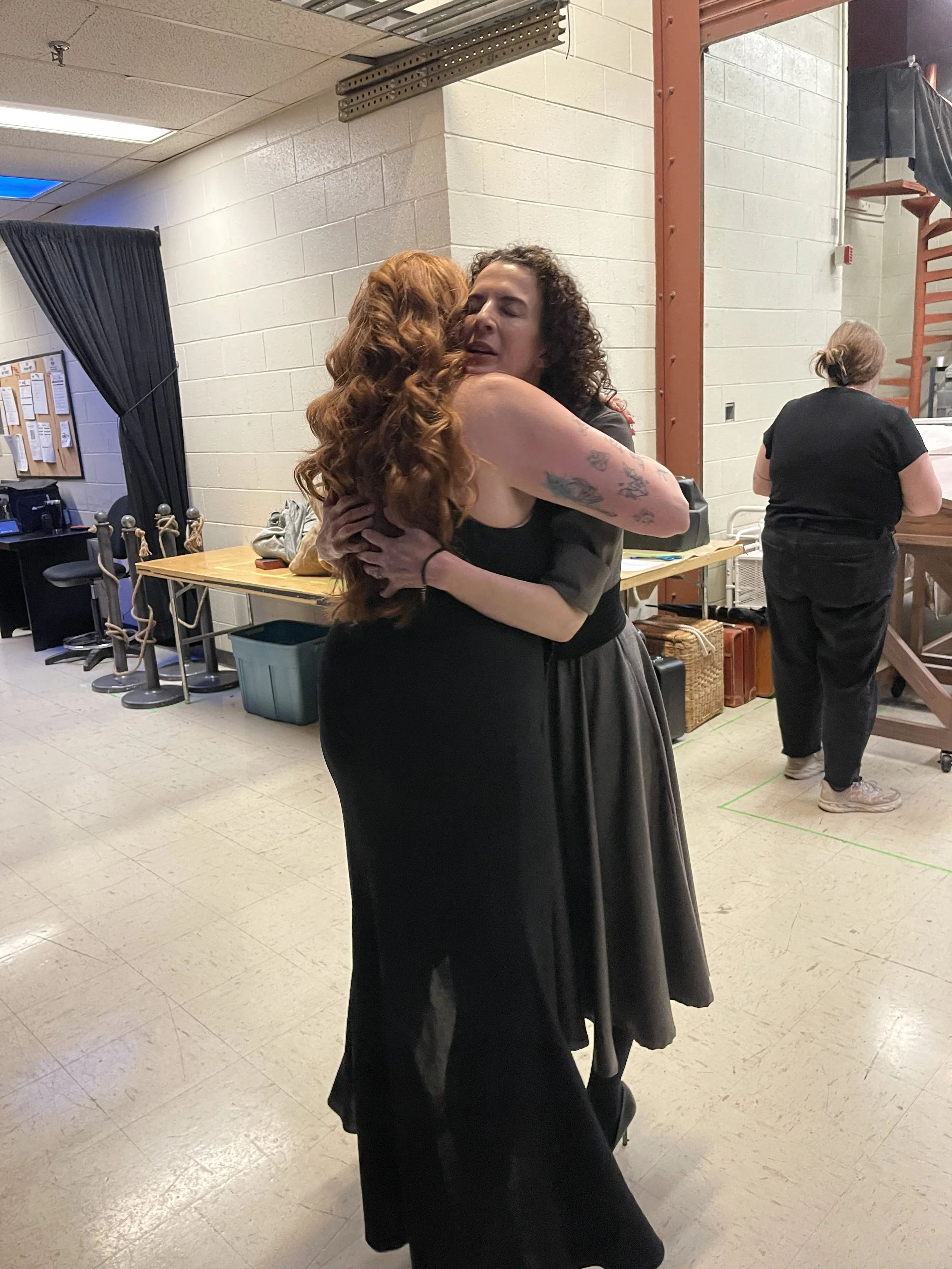 Two women are hugging each other in a room, one with curly black hair and the other with long, wavy red hair. One woman has tattoos on her arm.