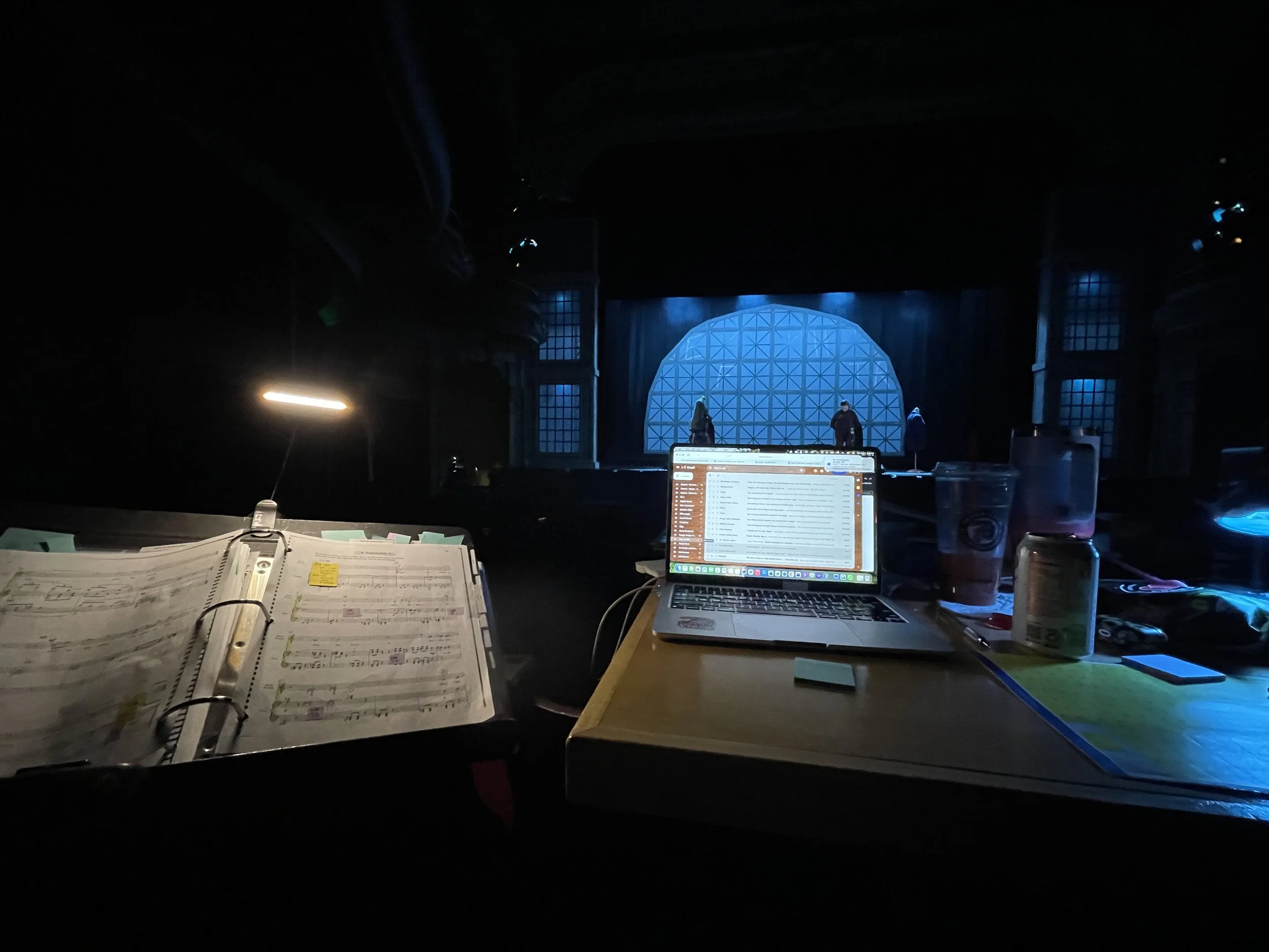 A dark stage with blue lighting and a large geometric blue backdrop, with three people on stage. In the foreground, a music sheet, laptop, and various items are visible on a table.