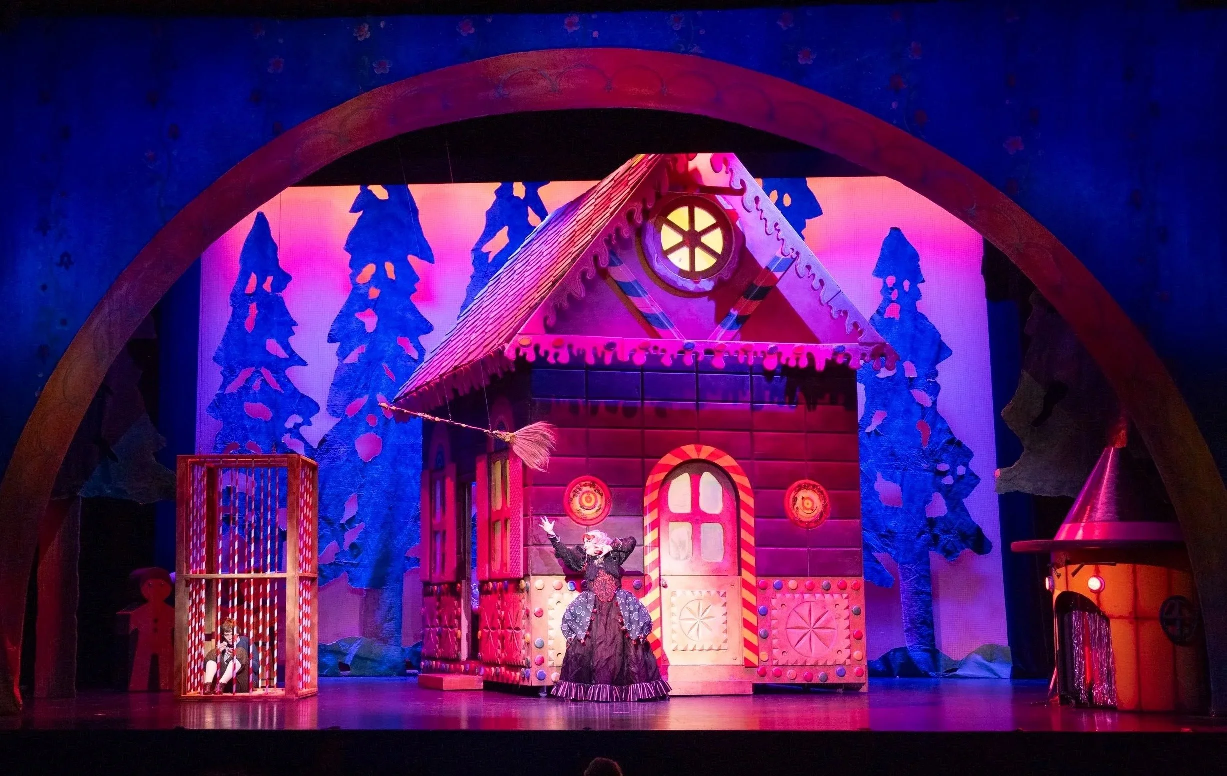 Theatrical set of a gingerbread house with colorful decorations, characters, and lights.