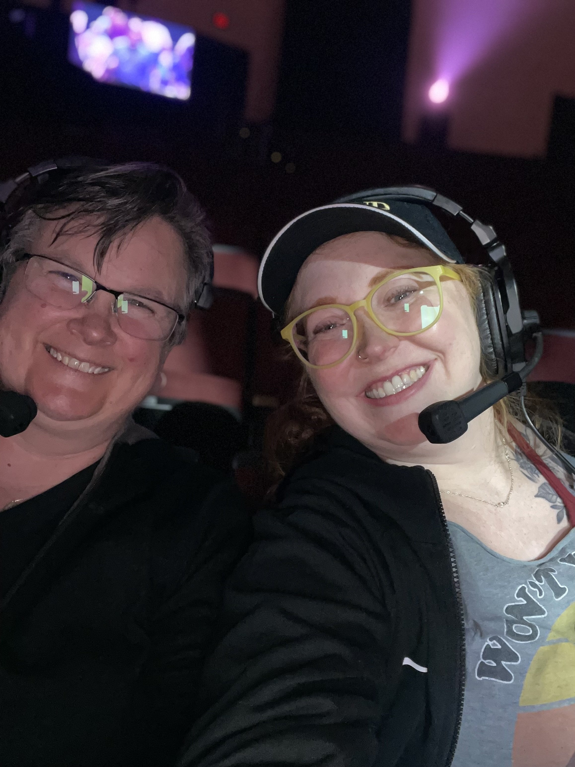 Two smiling people taking a selfie in a dark environment with a screen and purple light in the background, wearing headsets with microphones.