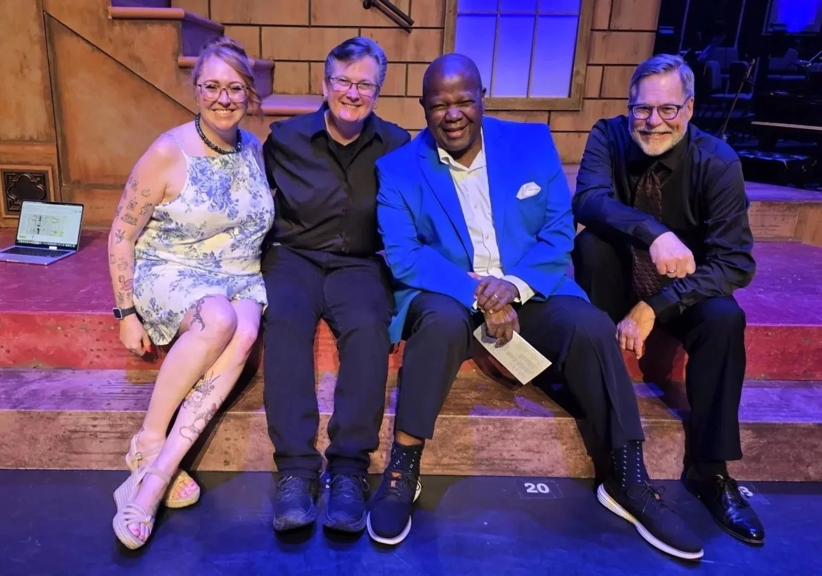 Four people sitting on a wooden stage, smiling at the camera. The woman on the left has tattoos and is wearing a floral dress, the second person is in black, the third is in a bright blue blazer, and the fourth is a man in black with glasses. There is a laptop on the stage to the left and a musical piano in the background.
