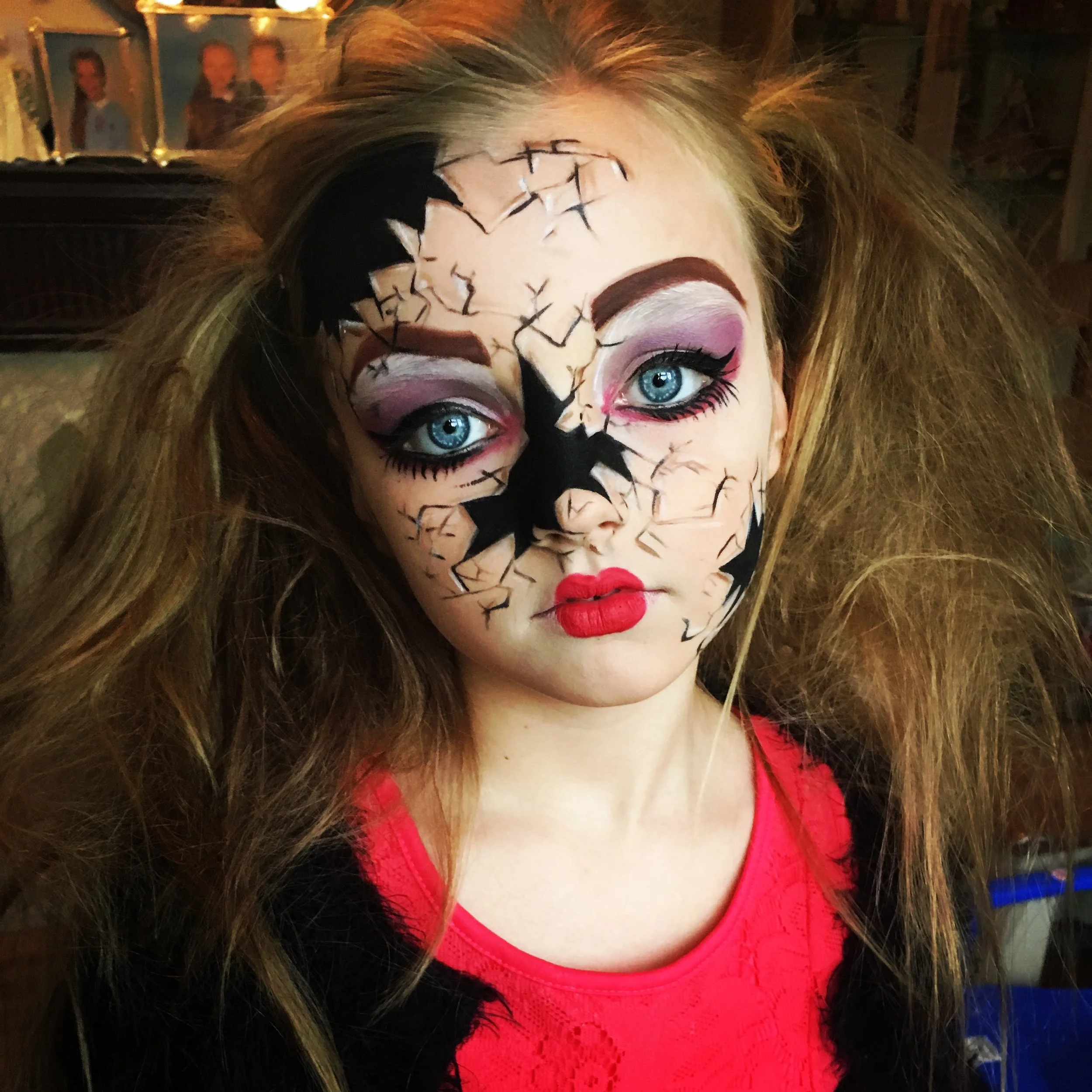 A woman with her face painted like a cracked porcelain doll, wearing a red top, with long curly hair, blue eyes, and makeup including pink eyeshadow and bright red lipstick.