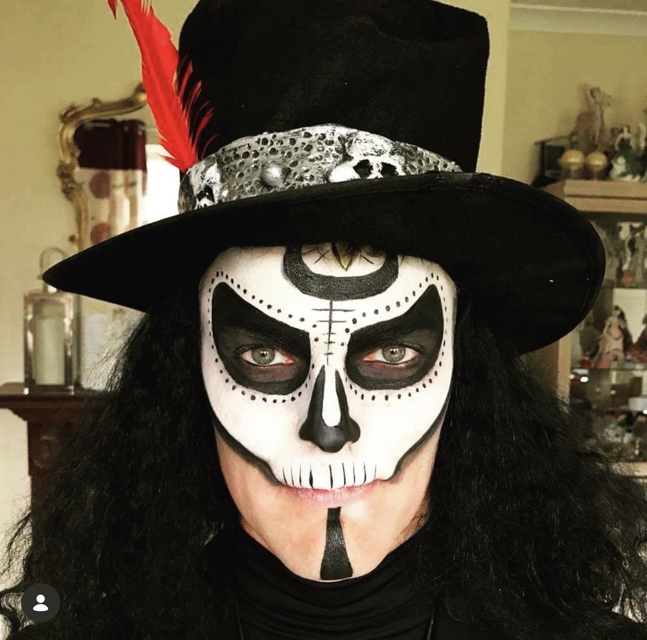 Person with intricate black and white skull face paint, wearing a black hat with a red feather and a decorative band, curly black hair, and a black shirt, indoors with shelves of figurines and decorative items in the background.