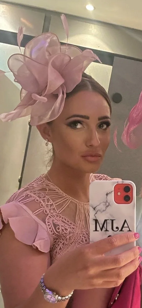 Woman taking a selfie with a smartphone in a room with ceiling lights, wearing a large pink floral hat and a pink lace dress.