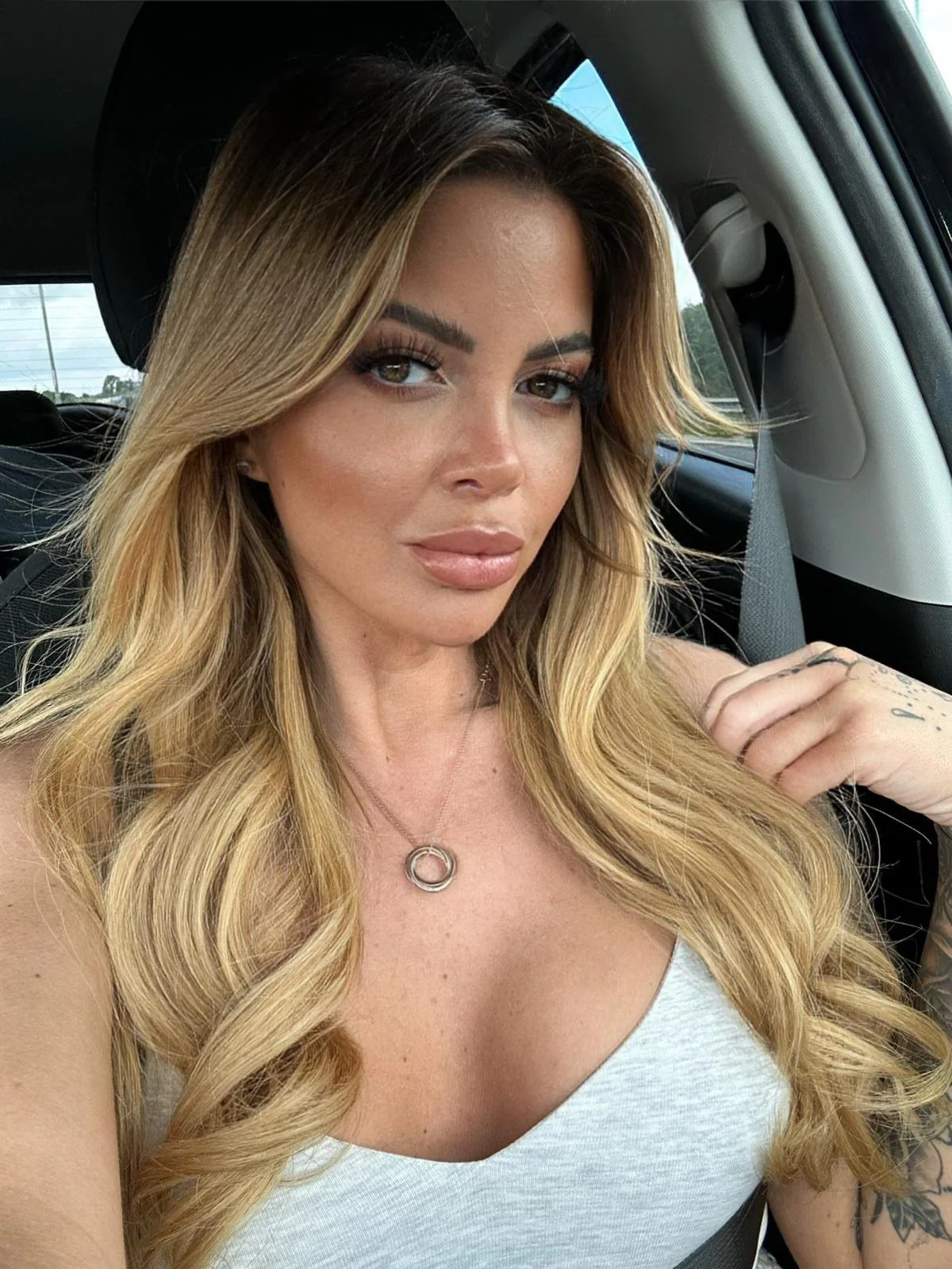 A woman with long blonde hair and makeup sitting in a car, wearing a gray tank top, a necklace with a circular pendant, and showing tattoos on her right arm.