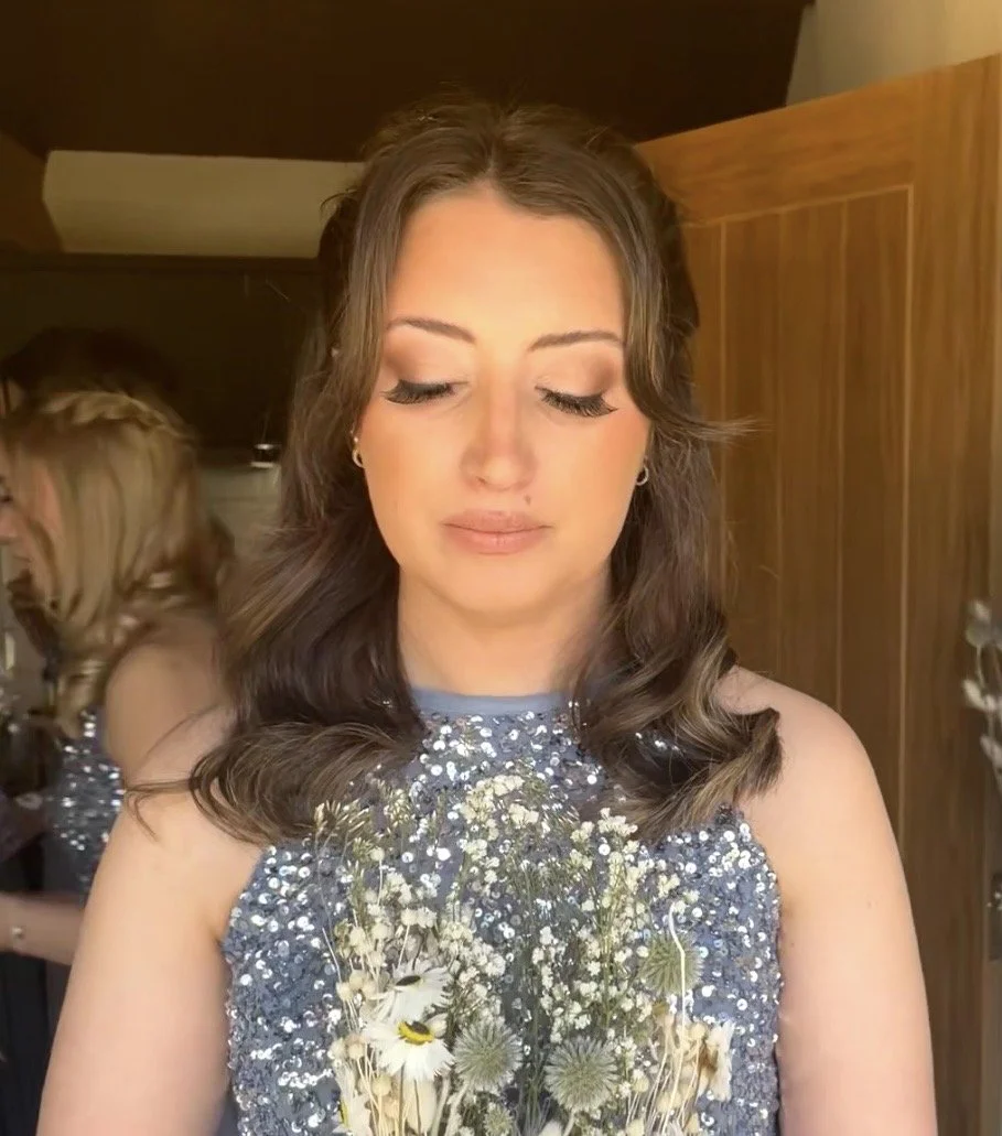 A woman with brown wavy hair and closed eyes holds a bouquet of wildflowers, wearing a sparkly sleeveless dress, in a room with wooden walls.