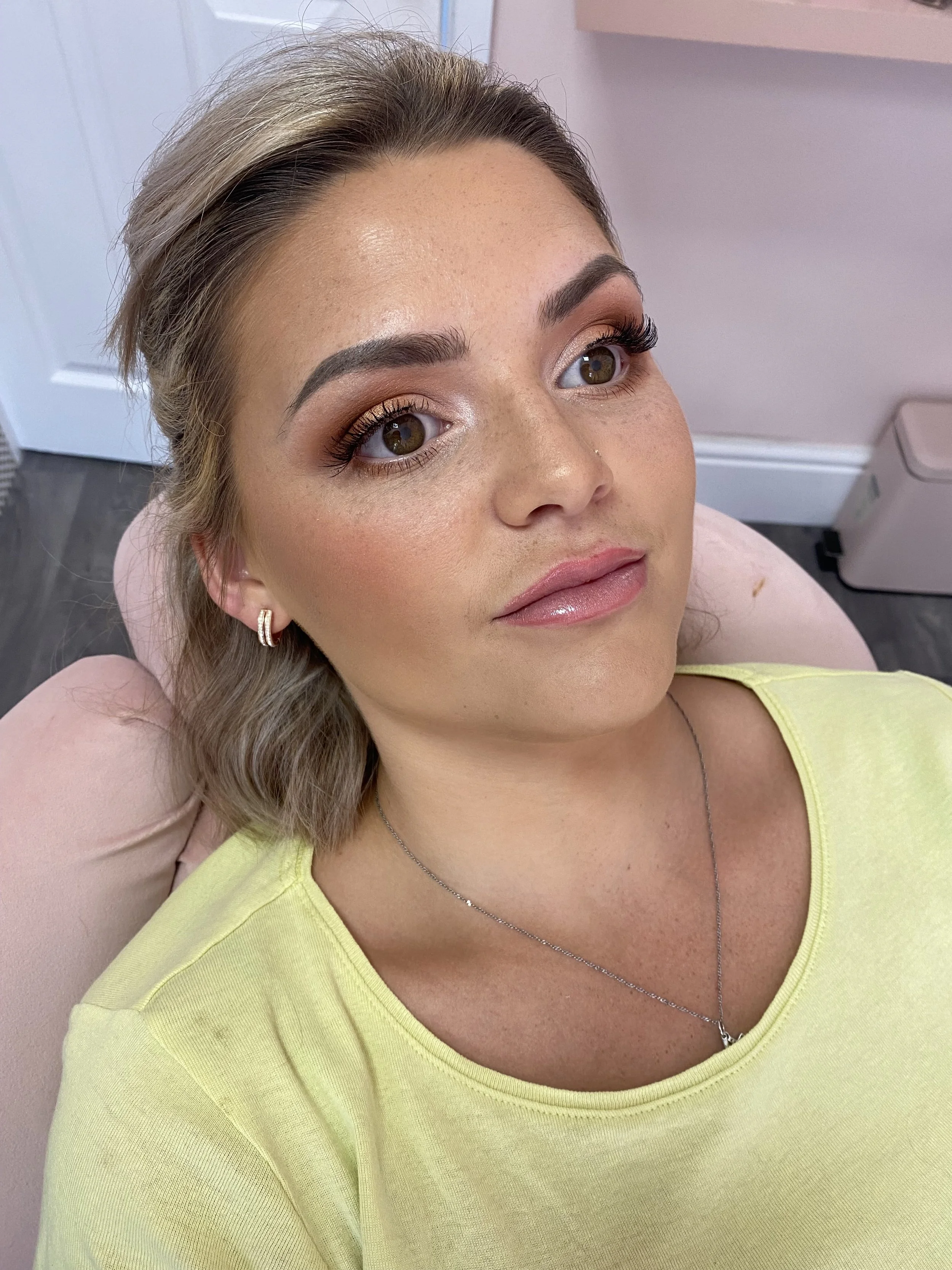 Close-up of a woman with styled blonde hair, brown eyes, makeup, wearing earrings, a yellow top, and a silver necklace, sitting in a room with pink walls.