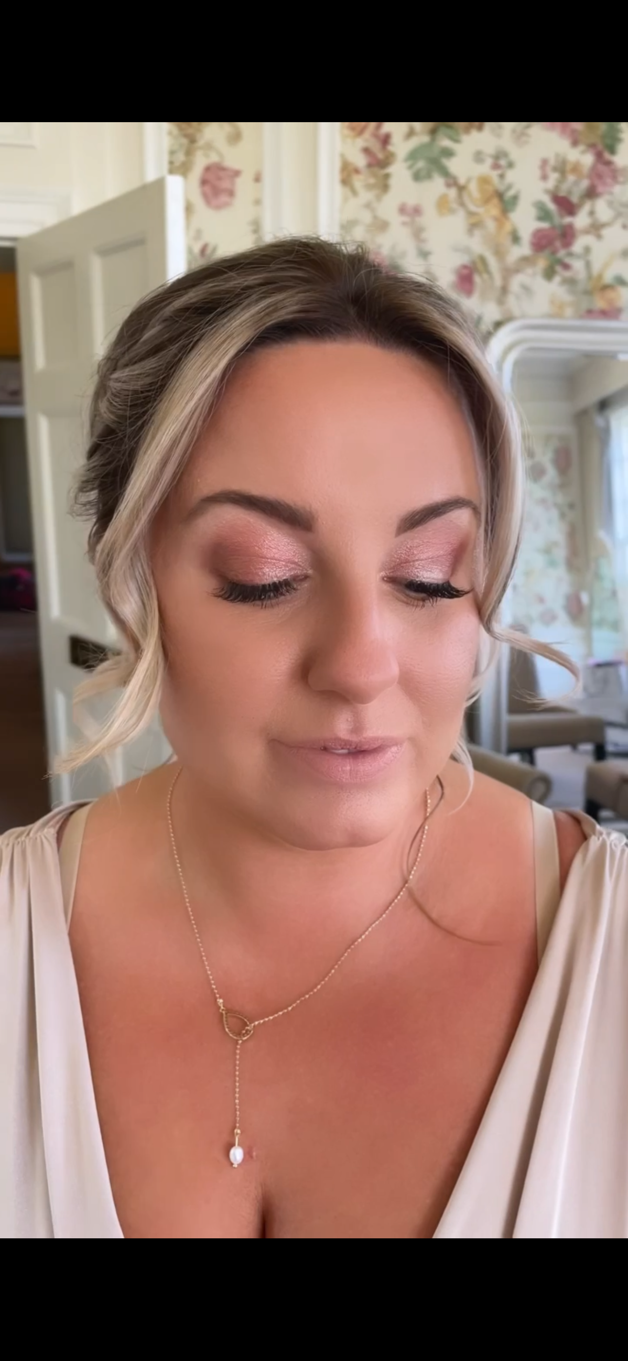 A woman with blonde hair styled in soft waves, wearing pink eyeshadow, long lashes, a light-colored dress, and a gold necklace with a pendant, smiling softly with her eyes closed in a room with floral wallpaper.