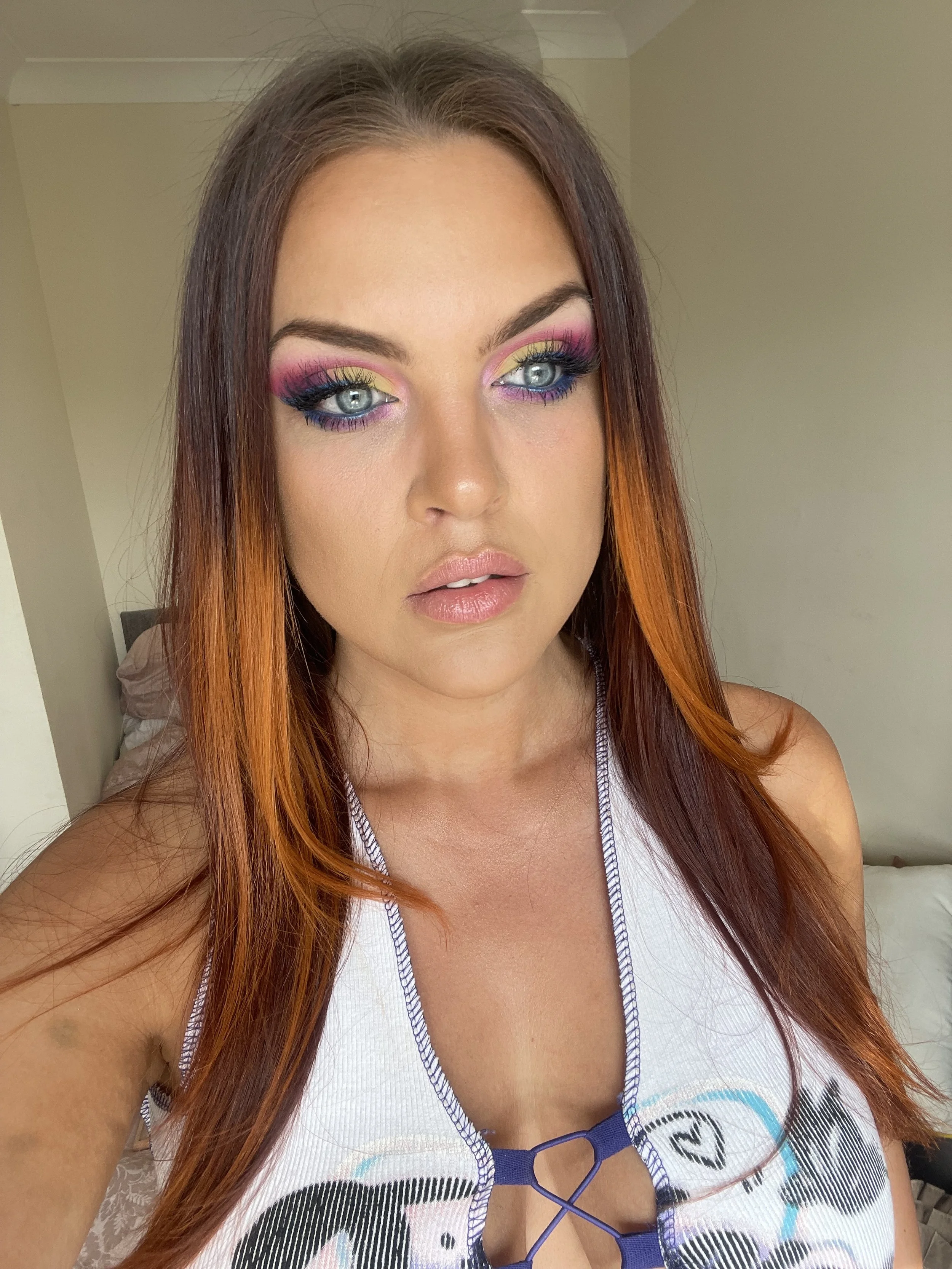 A young woman with long, wavy reddish hair and blue eyes taking a selfie. She has colorful eye makeup with pink, yellow, and purple shades, wearing a white tank top with a graphic design.