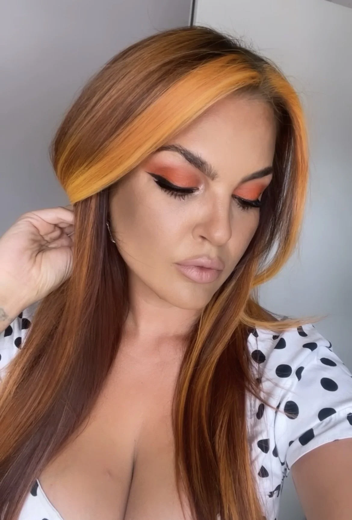 A woman with long, straight red hair and makeup with orange eyeshadow and winged eyeliner, wearing a white top with black polka dots, posing indoors.