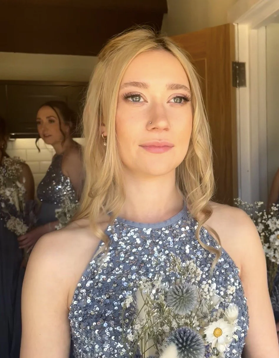 A young woman with blonde hair, blue eyes, and a nose piercing, wearing a sparkly, sequin dress, holding a bouquet of wildflowers, at a social gathering.