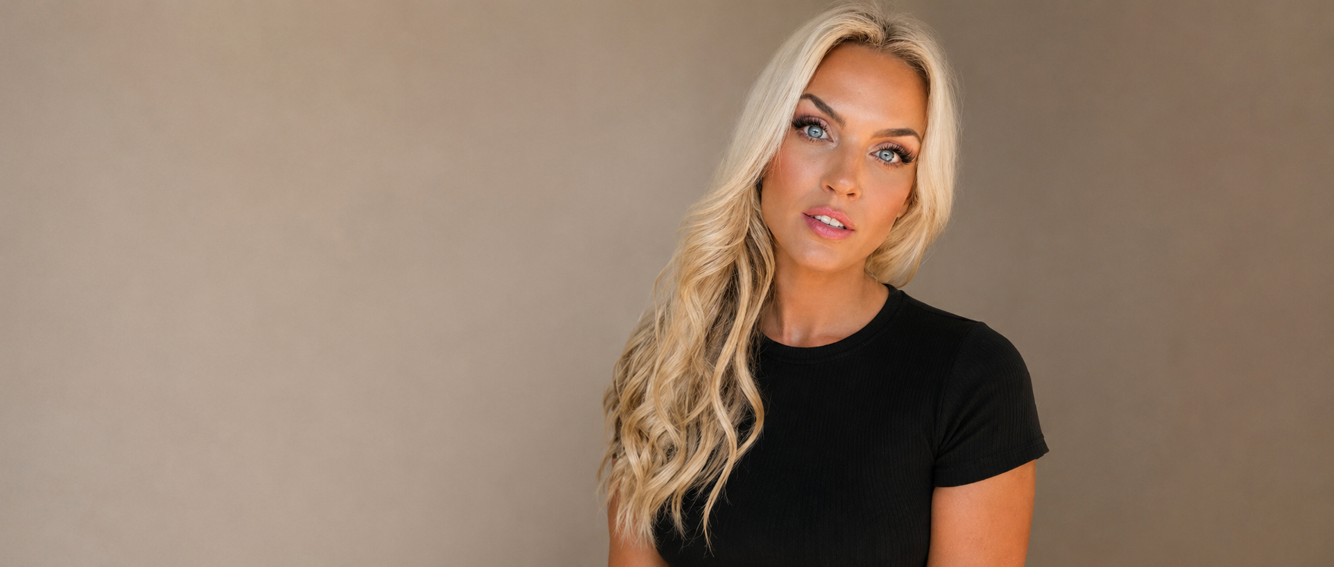 A woman with long blonde wavy hair and blue eyes, wearing a black t-shirt, looking directly at the camera against a plain beige background.